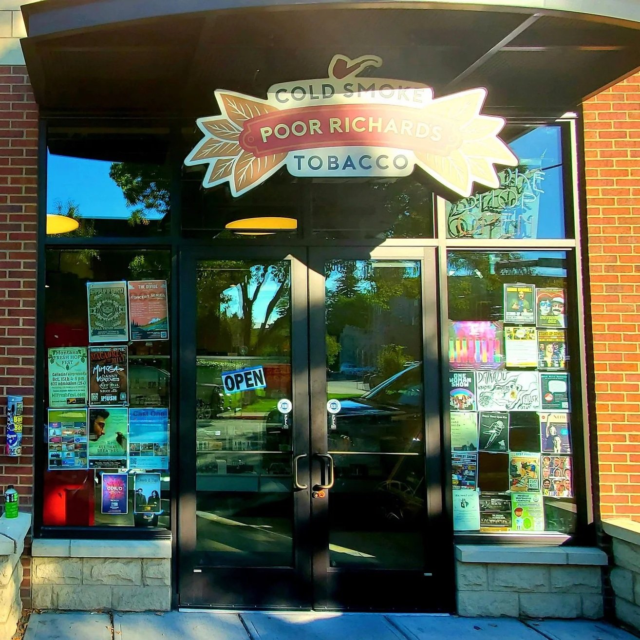 The storefront of Poor Richard's Tobacco, a tobacco shop, with a sign above the door reading 'Cold Smoke Poor Richard's Tobacco'. The glass windows display various posters and flyers. The door has an 'Open' sign. The building has a brick exterior.