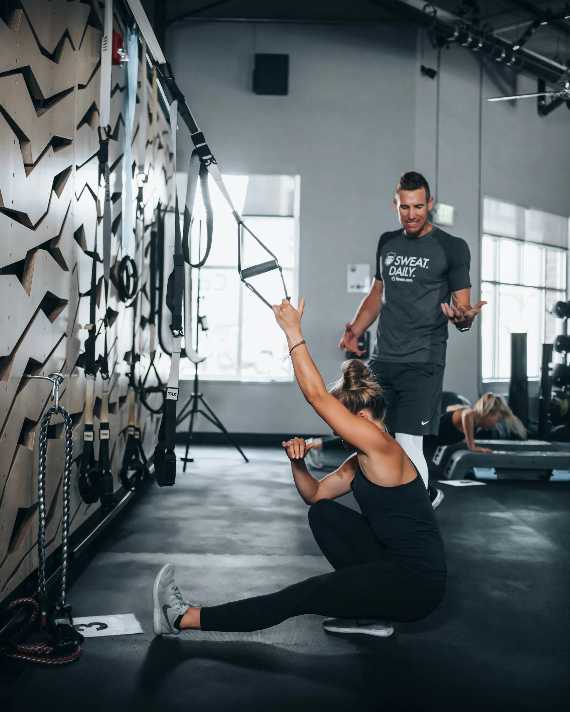 A woman is sitting on the gym floor performing a leg stretch with her legs extended and her arms holding her foot, while a male trainer in a gray shirt with the slogan 'SWEAT. DAILY.' watches and appears to offer guidance inside a fitness studio with suspension training straps on the wall.