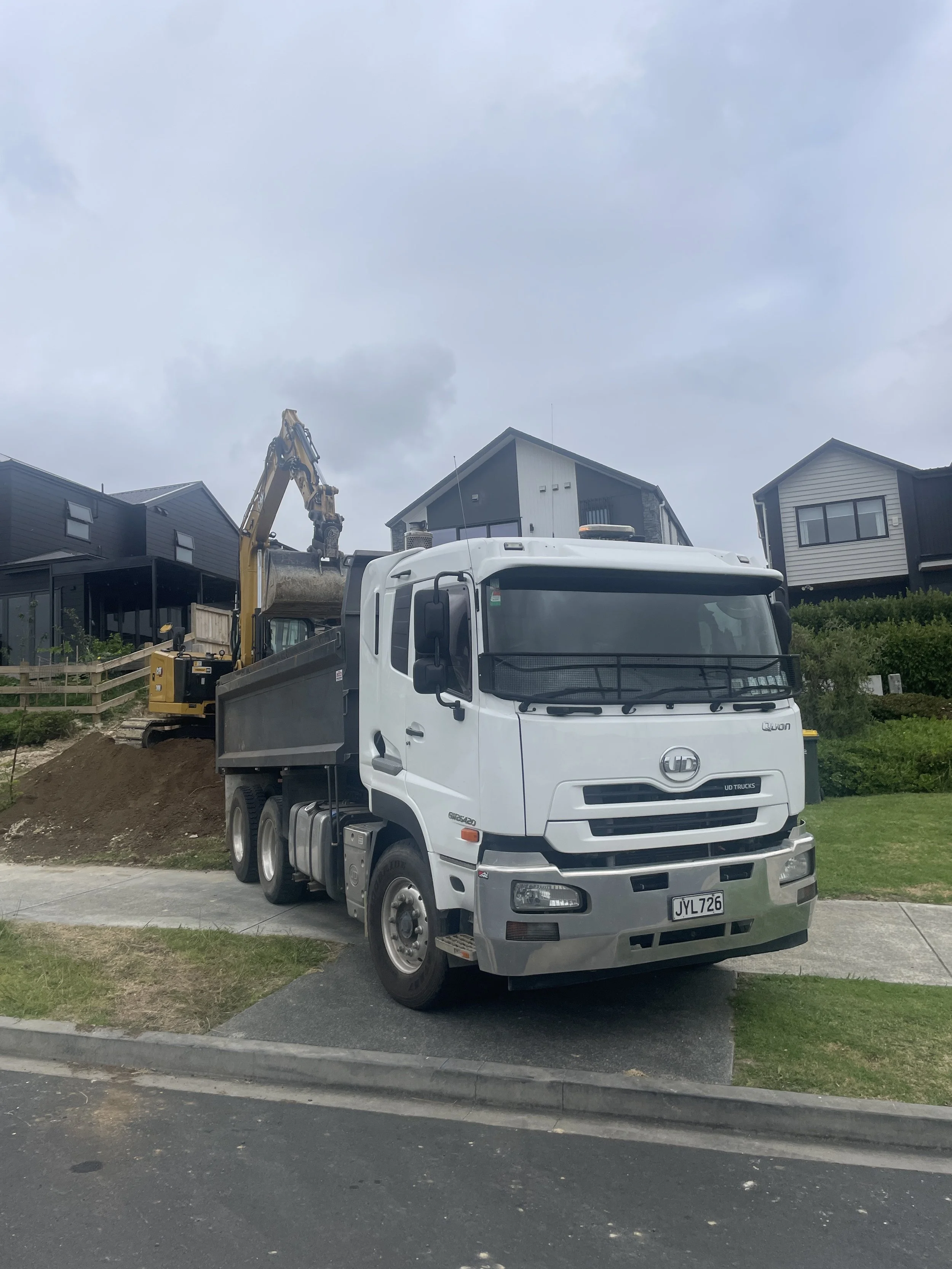 A white dump truck parked on the street with an excavator loading dirt into its bed, in front of modern houses on a cloudy day