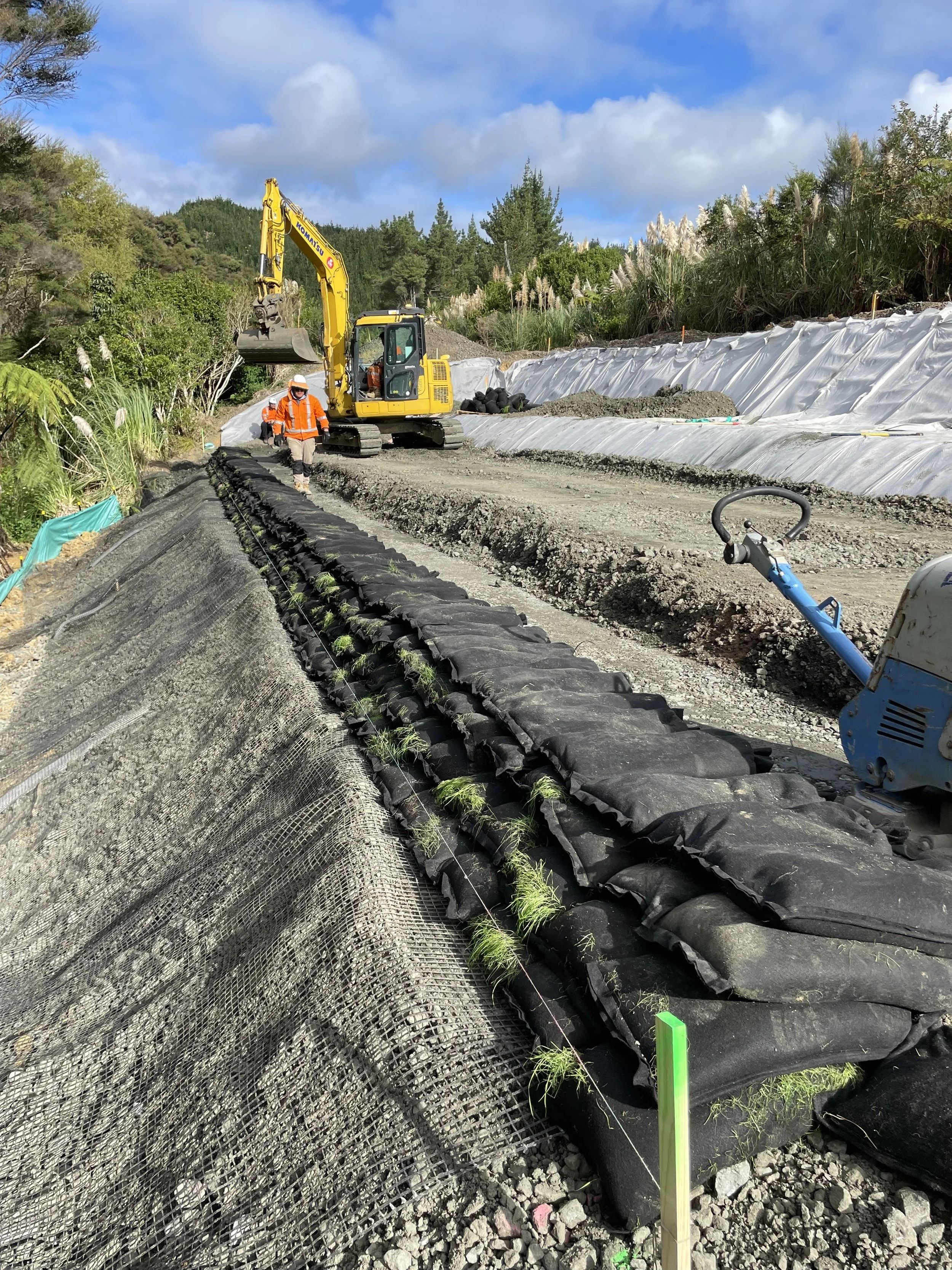 Construction workers planting grass on a hillside with an excavator and tools on a gravel road, surrounded by trees and a cloudy sky.