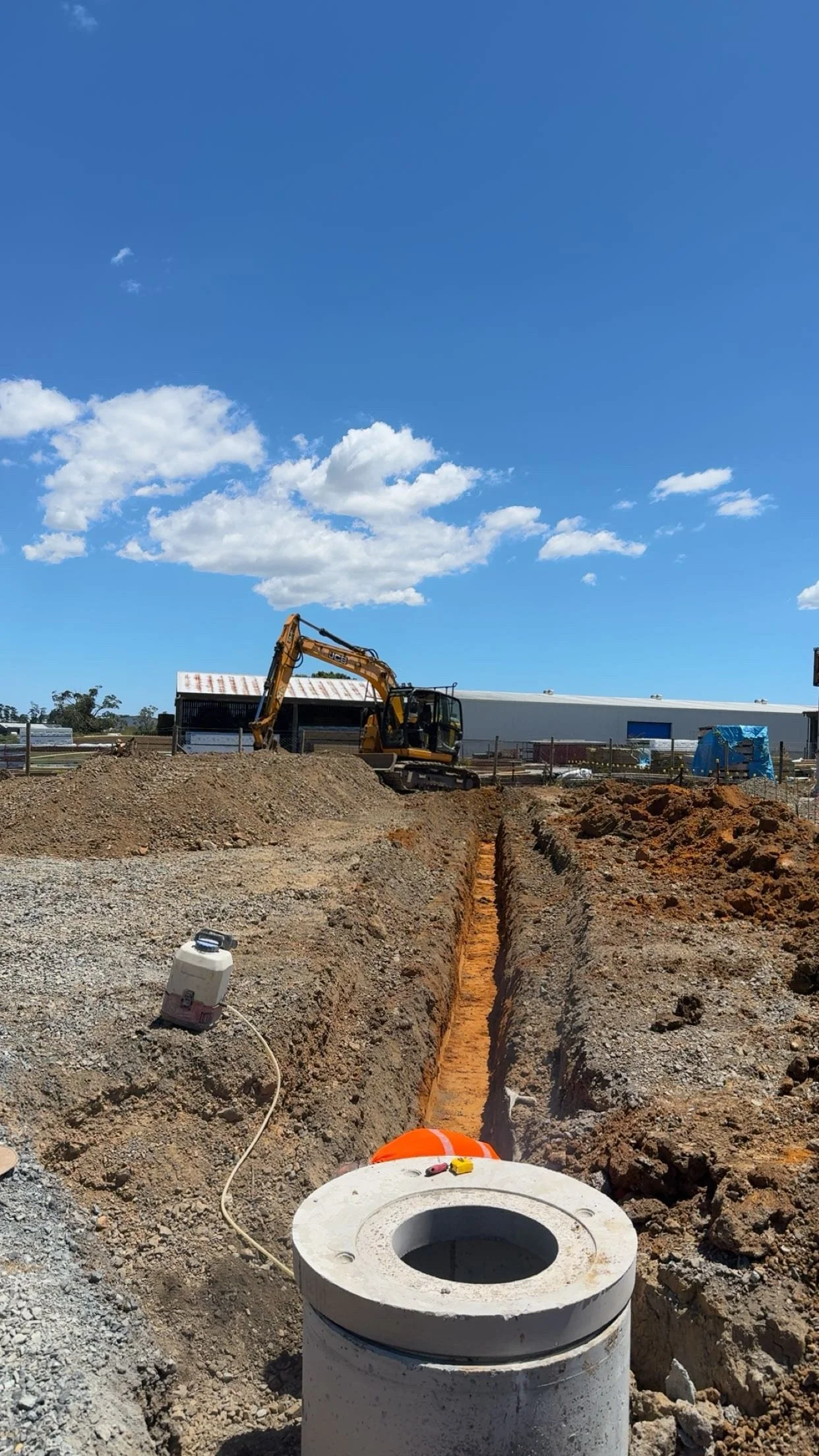 Construction site with a small excavator digging a trench, construction materials, and equipment in a partly cloudy sky background.