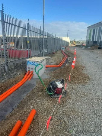 Construction site with orange pipes, a small generator, and a fenced area on a gravel surface under a clear blue sky.