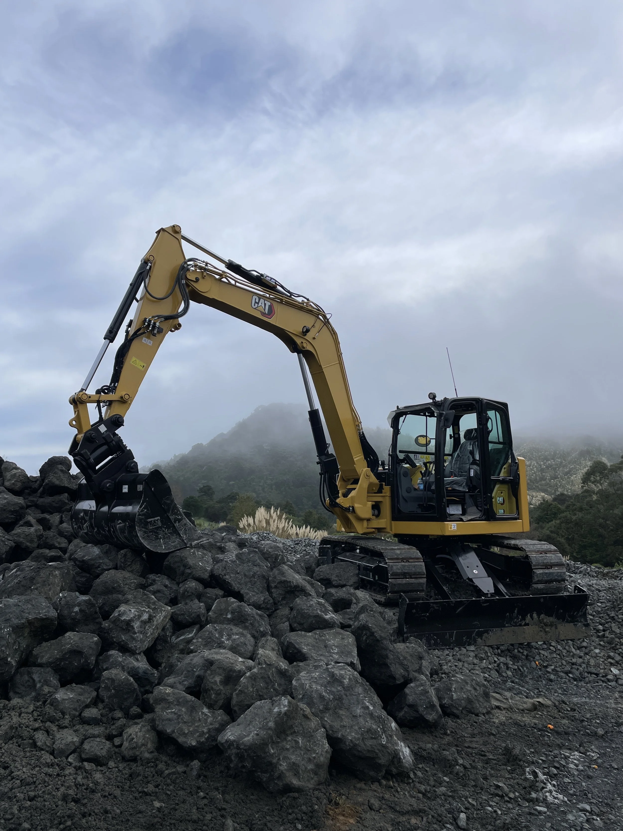Yellow Caterpillar construction excavator on rocky terrain with mountains in the background and overcast sky.