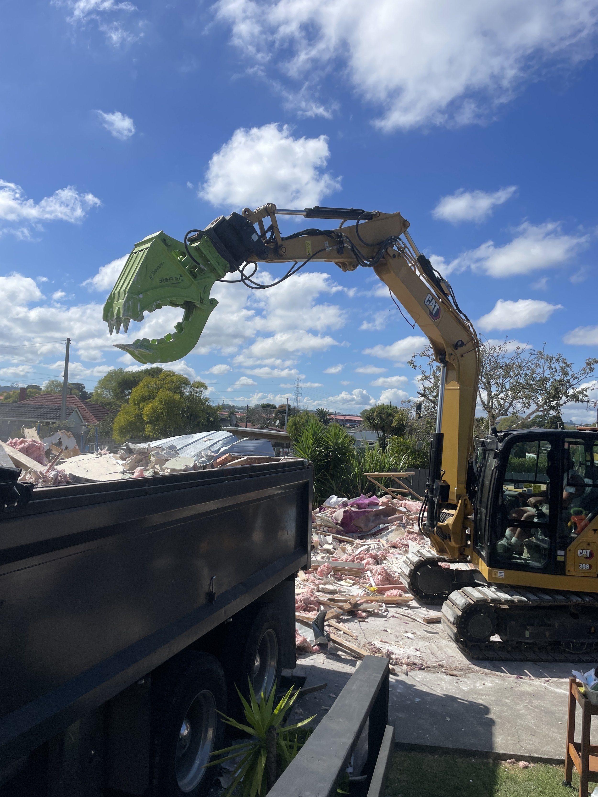A yellow excavator with a green claw lifting debris into a black dump truck on a clear day with blue sky and scattered clouds.