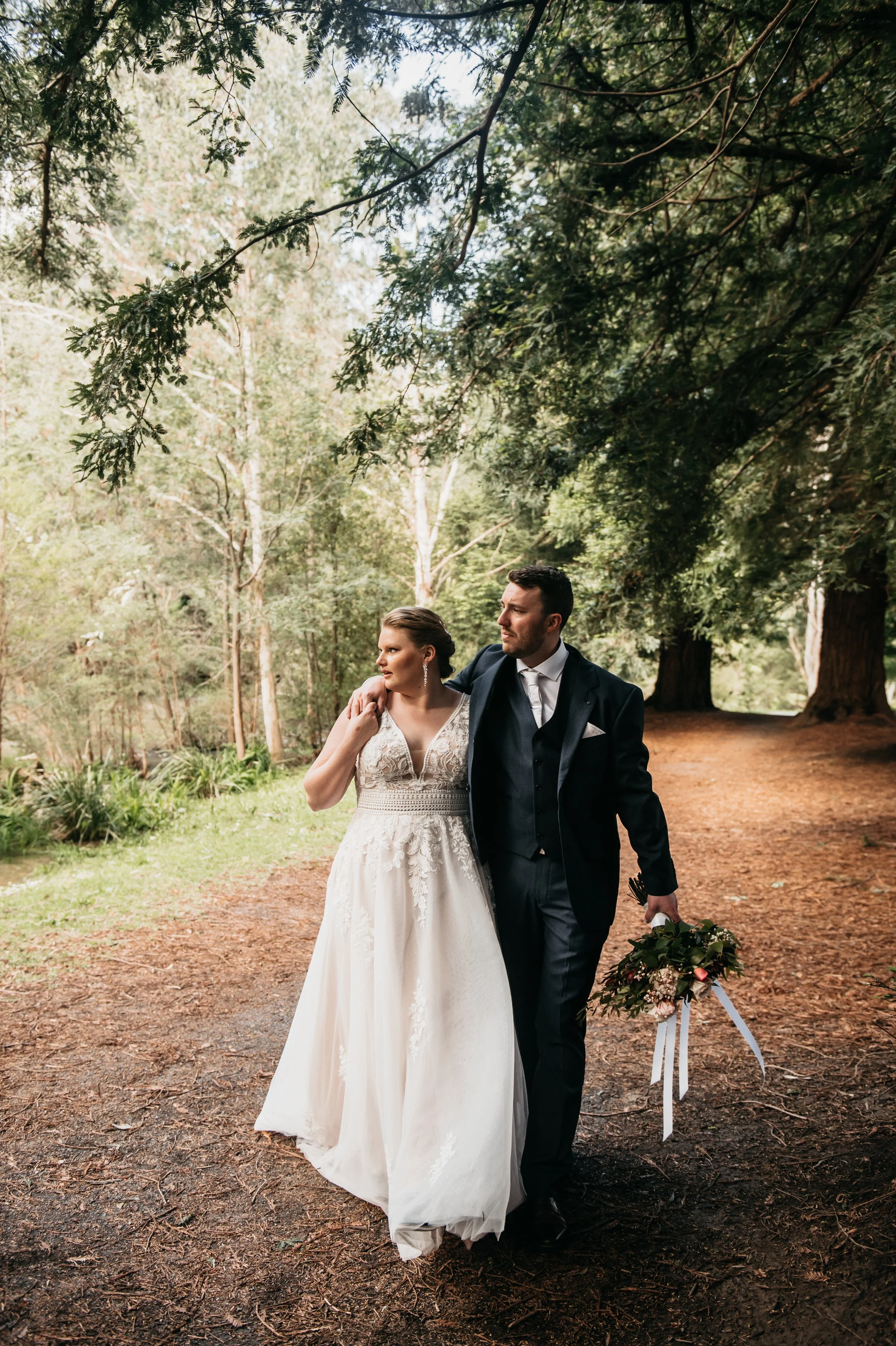 Bride and groom walking on a wooded path, surrounded by trees, during their wedding photo shoot, with the bride holding a bouquet.
