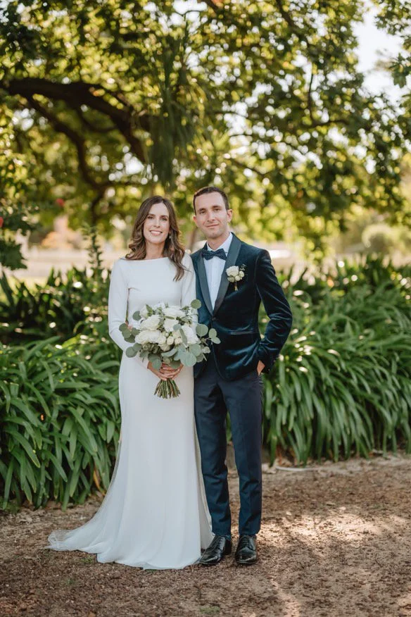 A bride and groom standing outdoors in a garden setting, smiling. The bride is wearing a long white wedding dress and holding a bouquet of white flowers and greenery. The groom is dressed in a dark blue tuxedo with a bow tie and boutonniere. There ar