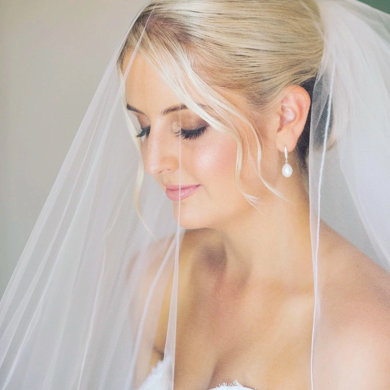 A bride with blonde hair wearing pearl earrings and a white veil, looking down softly with makeup and her hair styled elegantly.