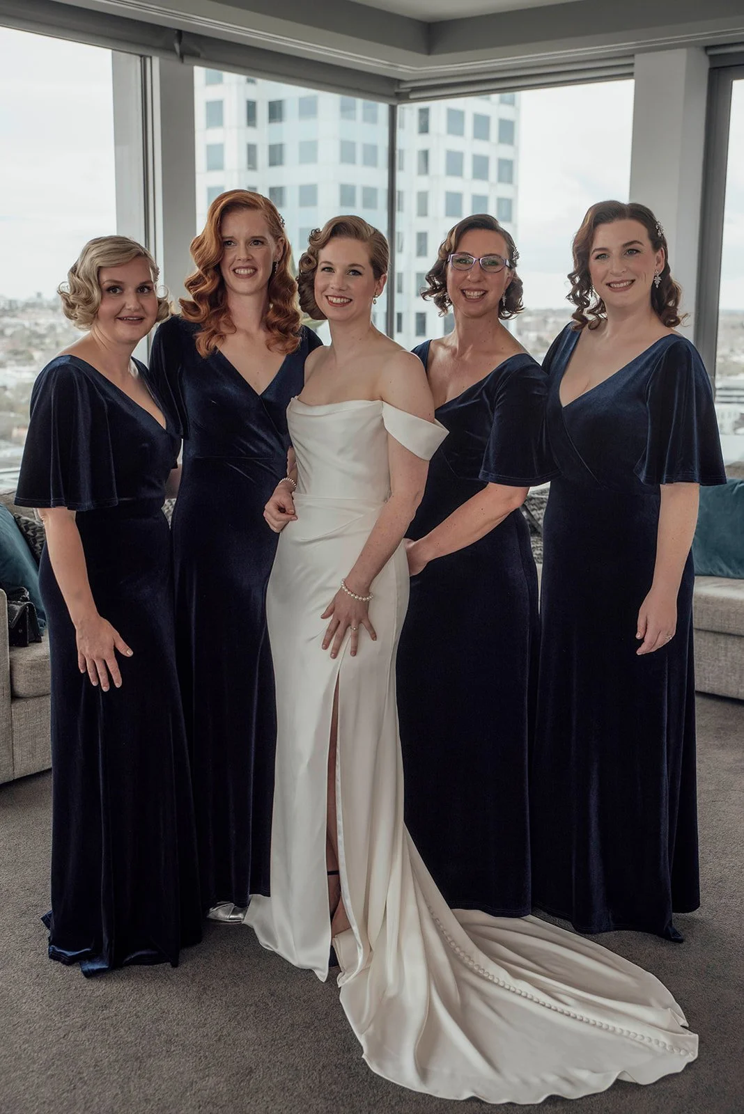 A bride in a white wedding dress with off-the-shoulder sleeves and a slit, standing with five bridesmaids in matching navy velvet dresses with flutter sleeves, inside a modern apartment with large windows and a city view.