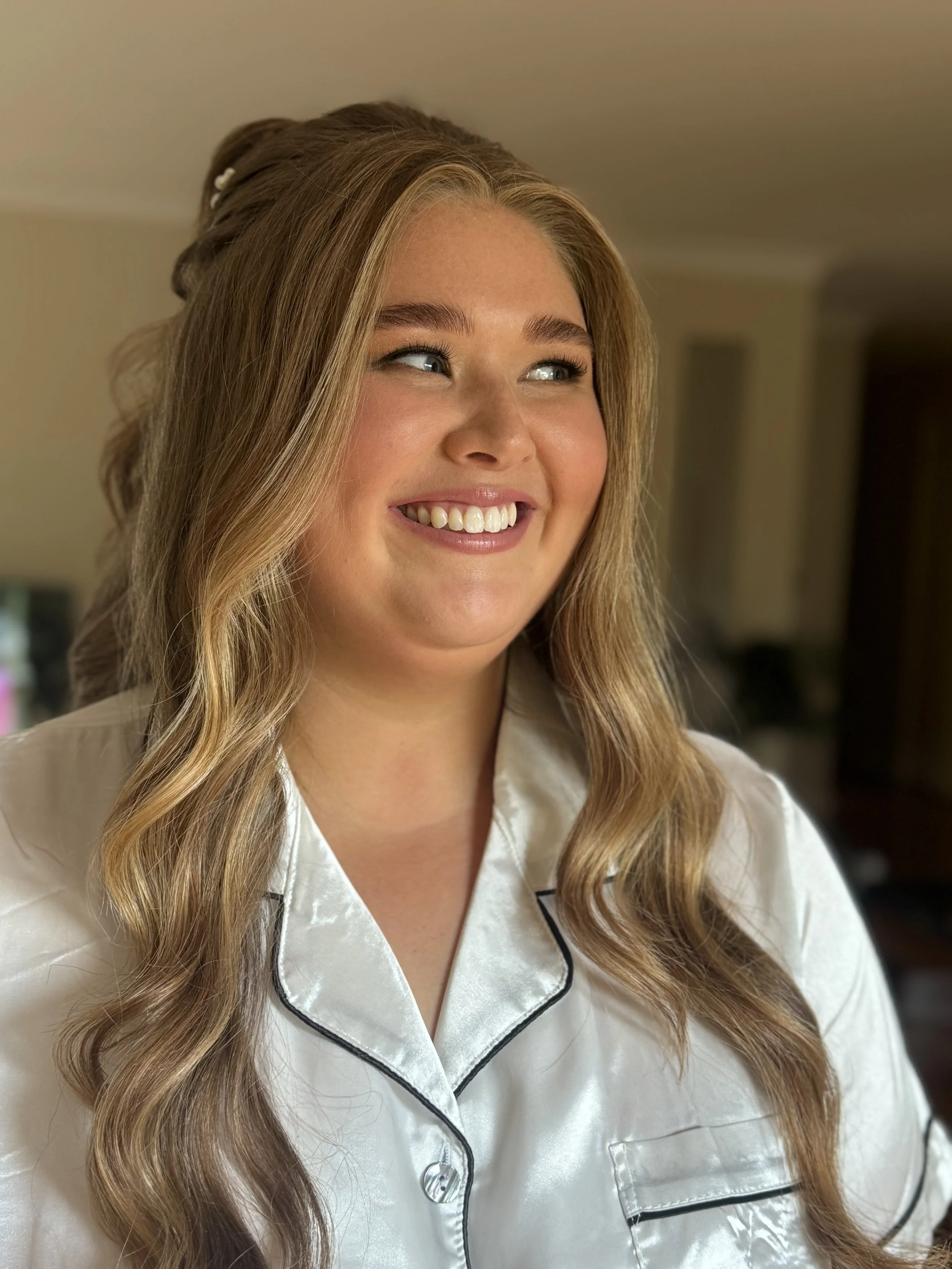 A smiling woman with long, wavy blonde hair wearing white satin pajamas with black piping, inside a home.