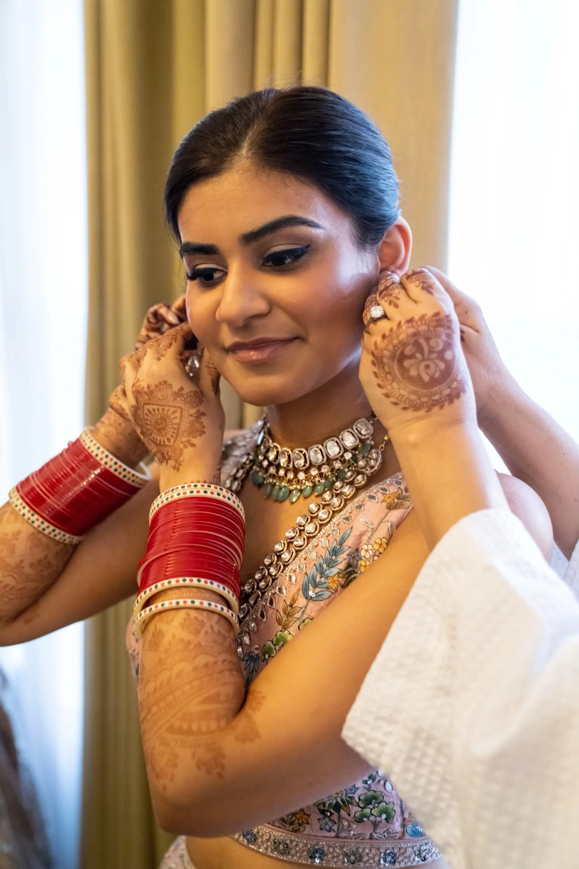 A woman dressed in traditional Indian attire, wearing jewelry and red bangles, is being helped to put on earrings, with hands adjusting her earrings.