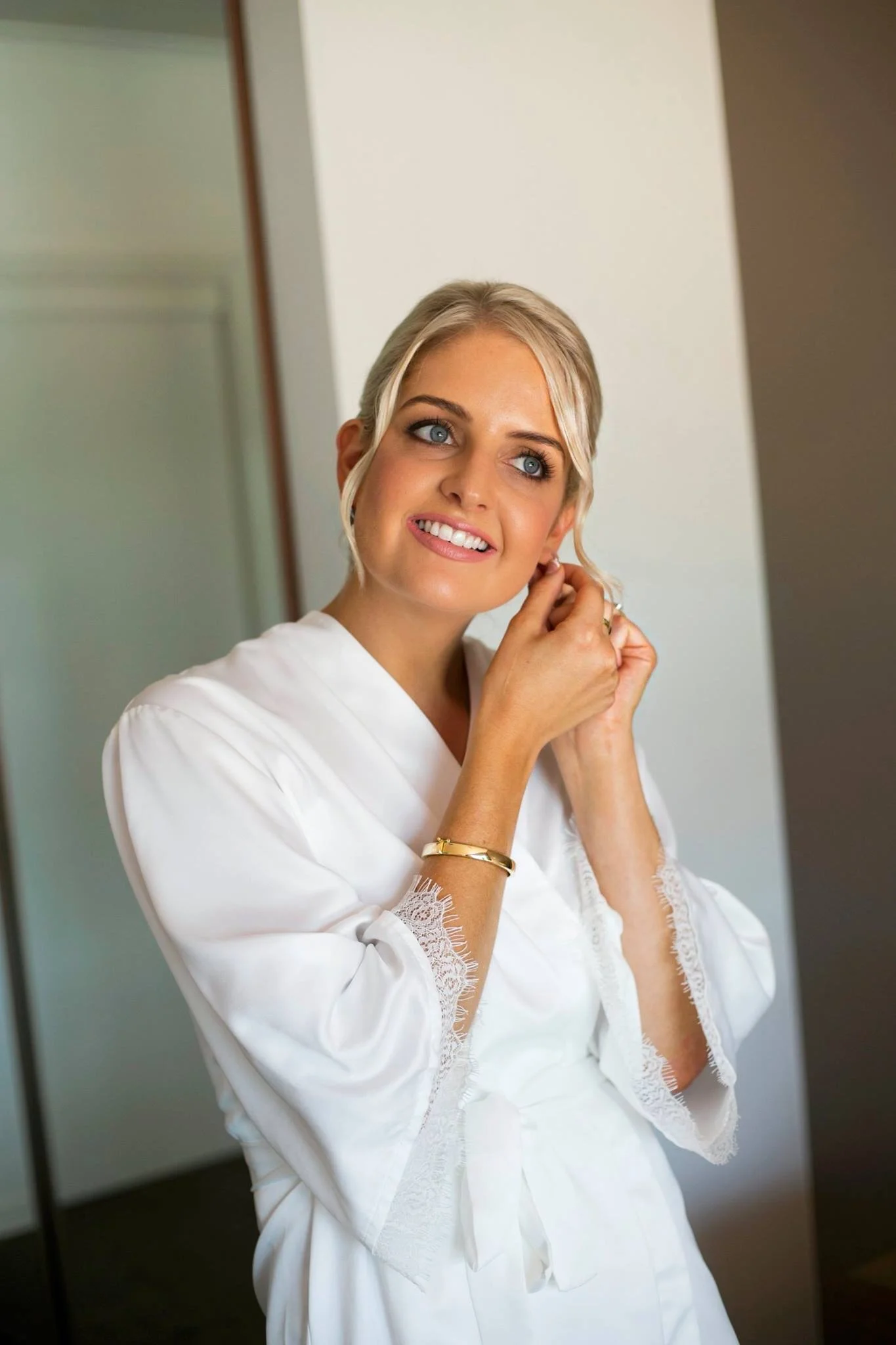 A young woman with blonde hair and blue eyes is putting on earrings. She is wearing a white satin robe with lace detailing, and is smiling while looking slightly to her left.