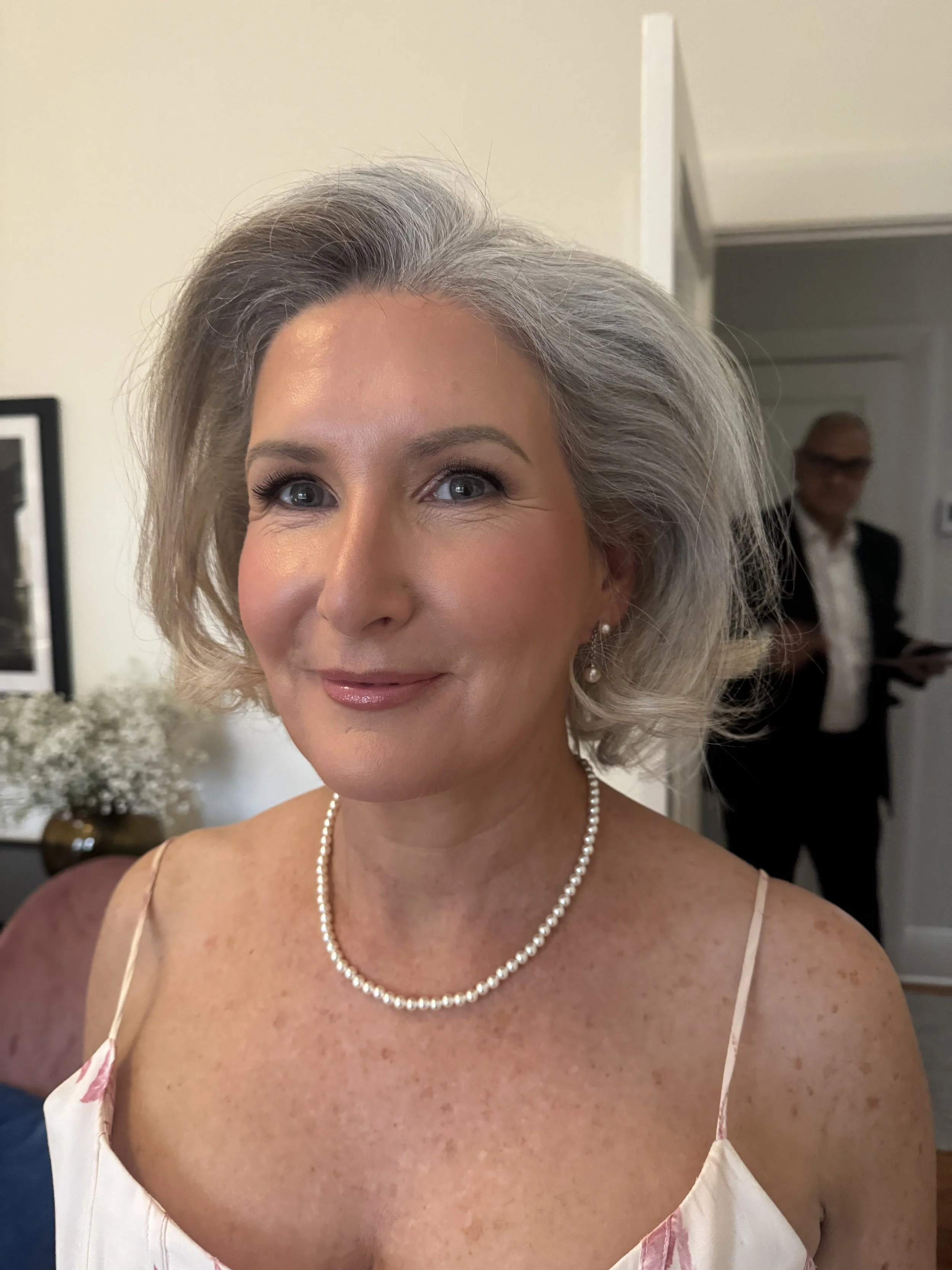 Natural makeup and hair on a mature bride getting ready for her wedding in Elsternwick. She is sitting in a house with flowers and a male in the background.
