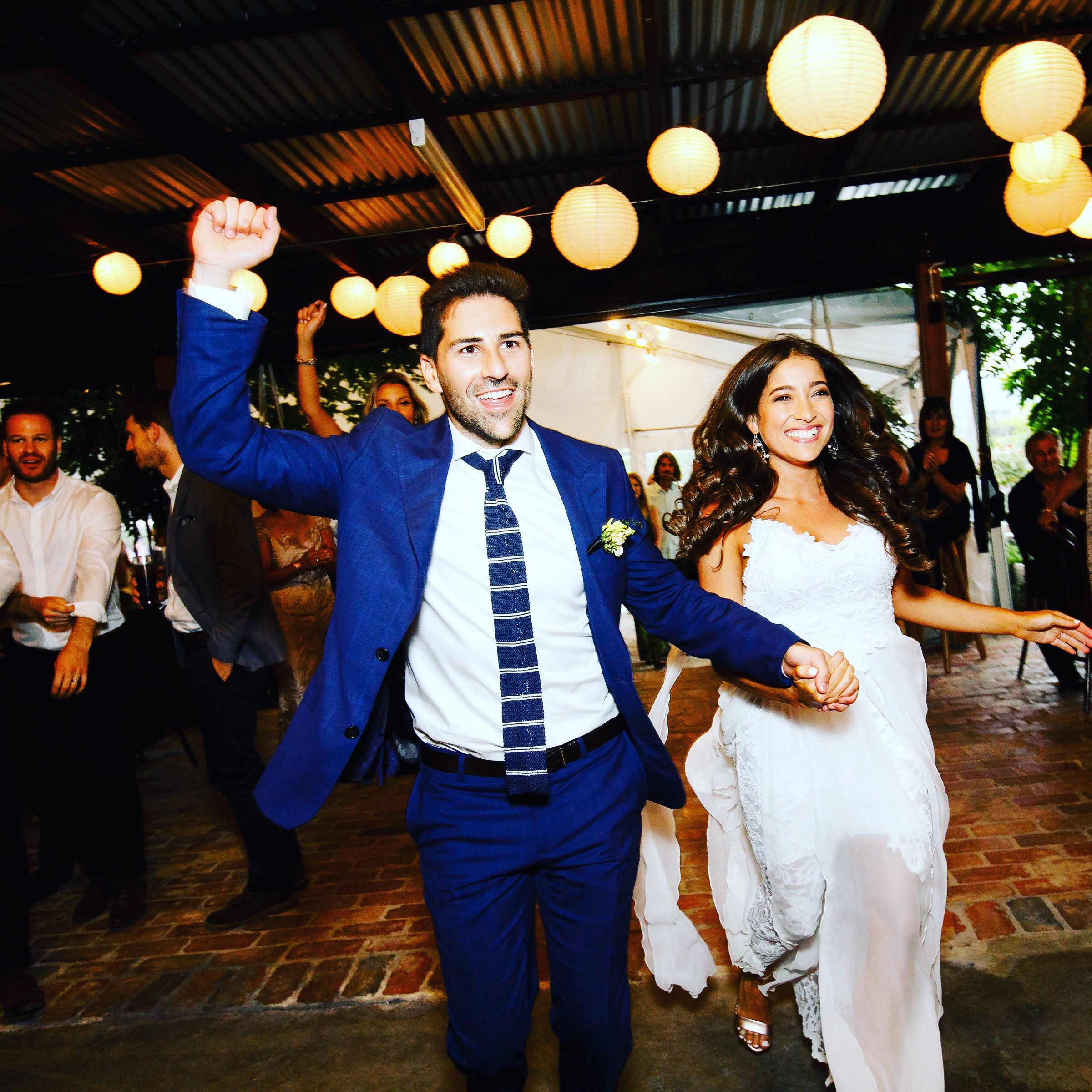 A newlywed couple holding hands and dancing at their wedding reception, with wedding guests in the background under hanging paper lanterns.