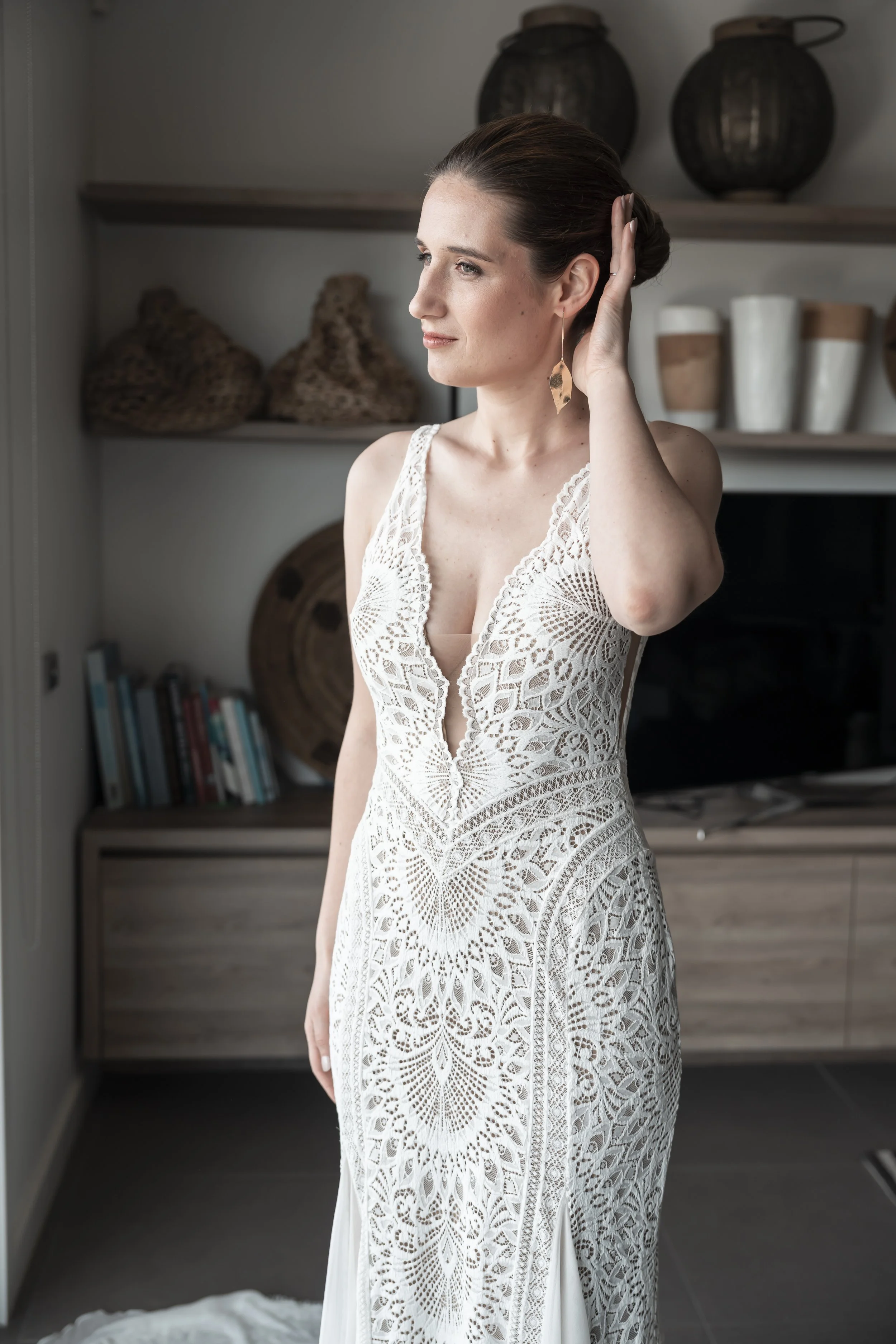 A woman in a white lace dress looking to the side, holding her ear, standing in a room with shelves and decorative items in the background.