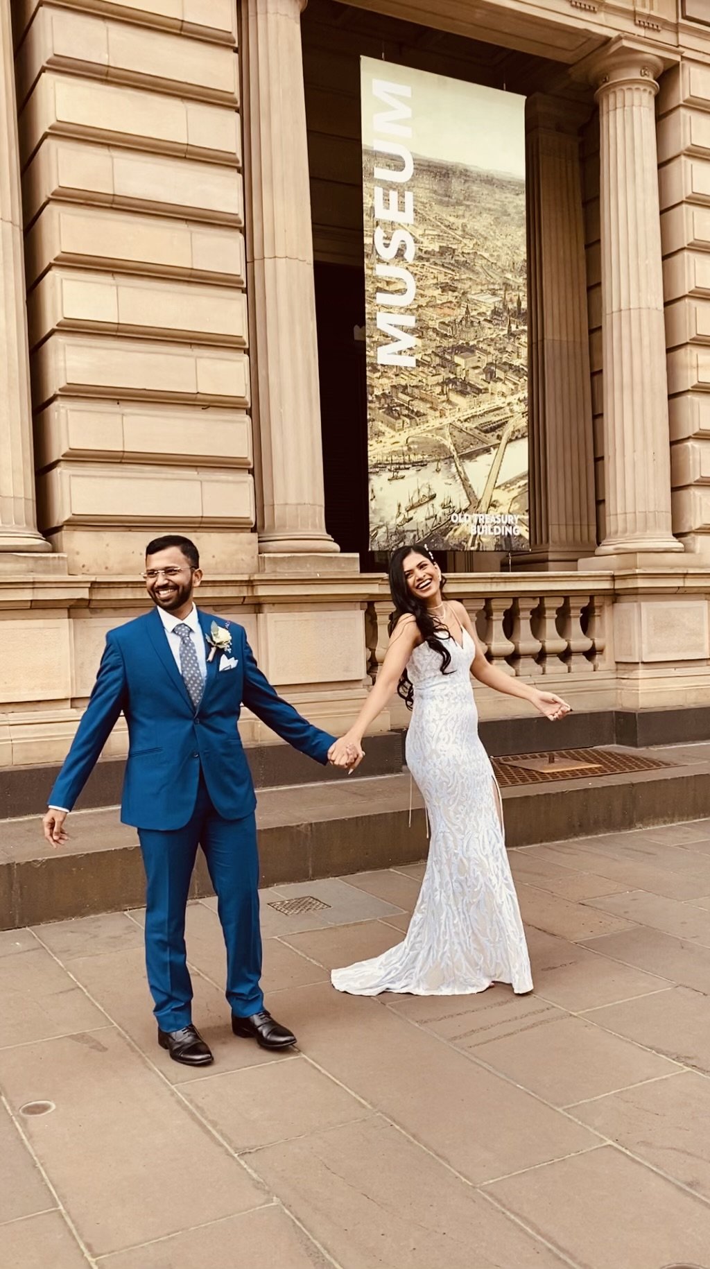A couple in wedding attire standing outside a building with large columns, holding hands and smiling. The man is in a blue suit and the woman in a white dress. A large museum banner is visible in the background.