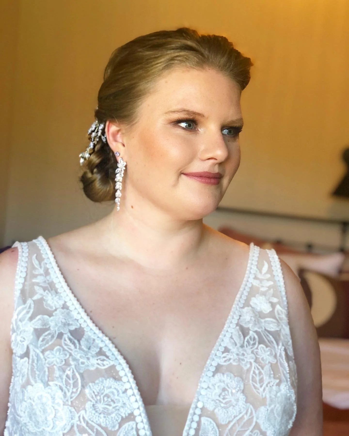 Close-up of a bride with light skin, blue eyes, and a gentle smile, wearing a lace wedding dress with floral embroidery and pearl embellishments, and long dangling earrings with gemstones. Her hair is styled in an elegant updo.