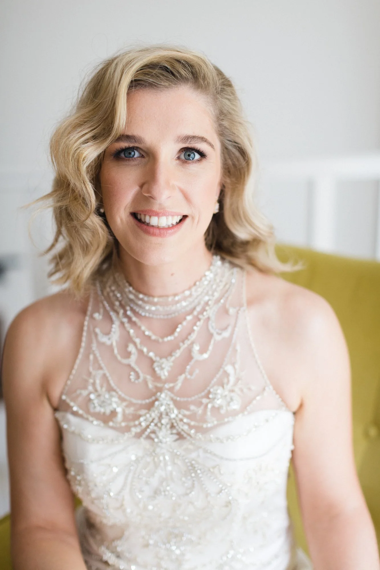 A woman with blonde wavy hair, blue eyes, and fair skin, smiling, wearing a white wedding dress with intricate beadwork and pearl necklaces, seated on a yellow chair in a bright room.