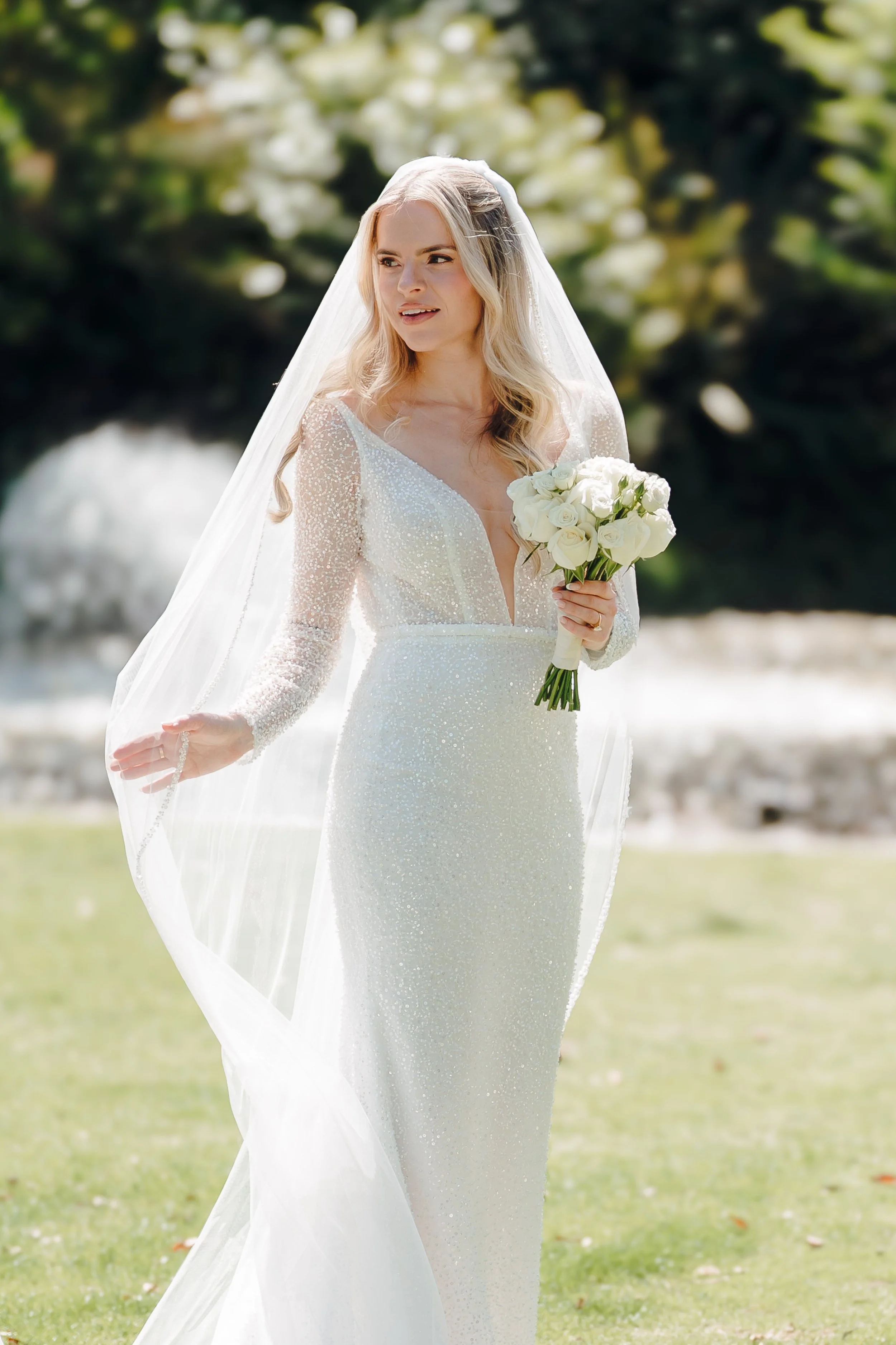 A bride in a white, long-sleeved, sequined wedding dress holding a bouquet of white roses outdoors with trees and grass in the background.