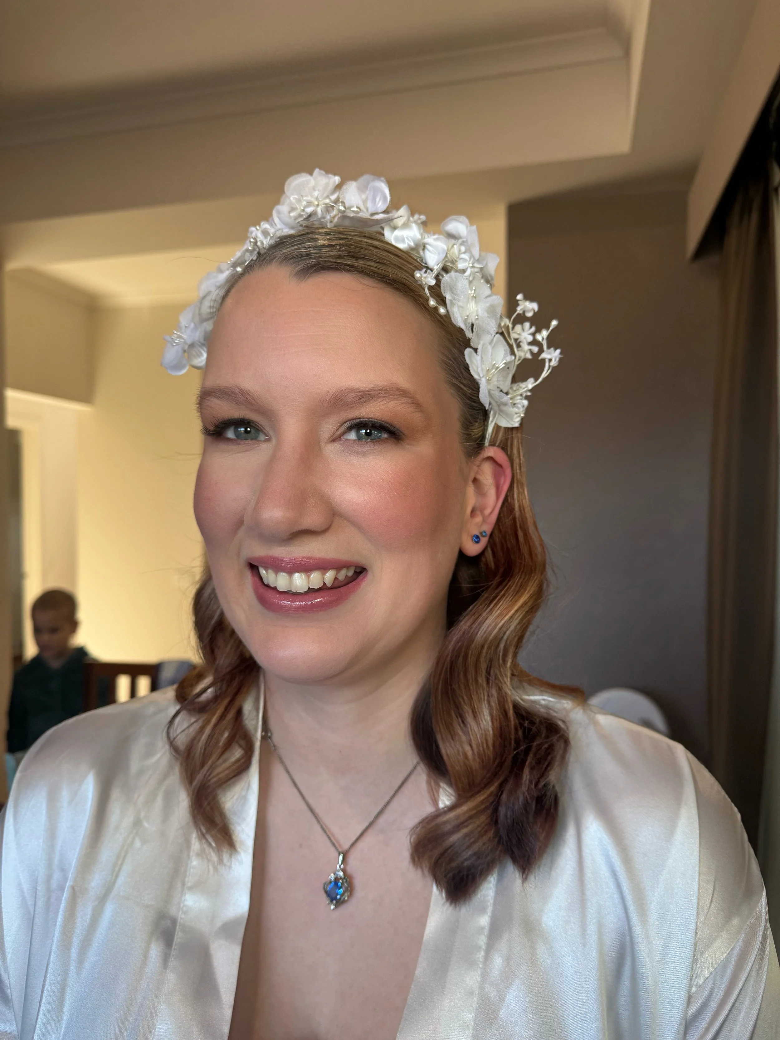 A woman with wavy light brown hair, wearing a floral headband, pearl earrings, a necklace with a blue pendant, and a cream-colored satin robe, smiling in a room with beige walls and a window with dark curtains. In the background, a young boy is sitti