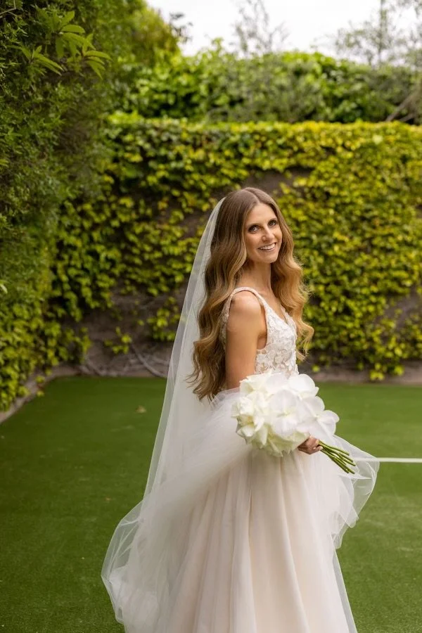 A bride in a white wedding gown holding a bouquet of white flowers, standing on a grassy area outdoors with a hedge background.