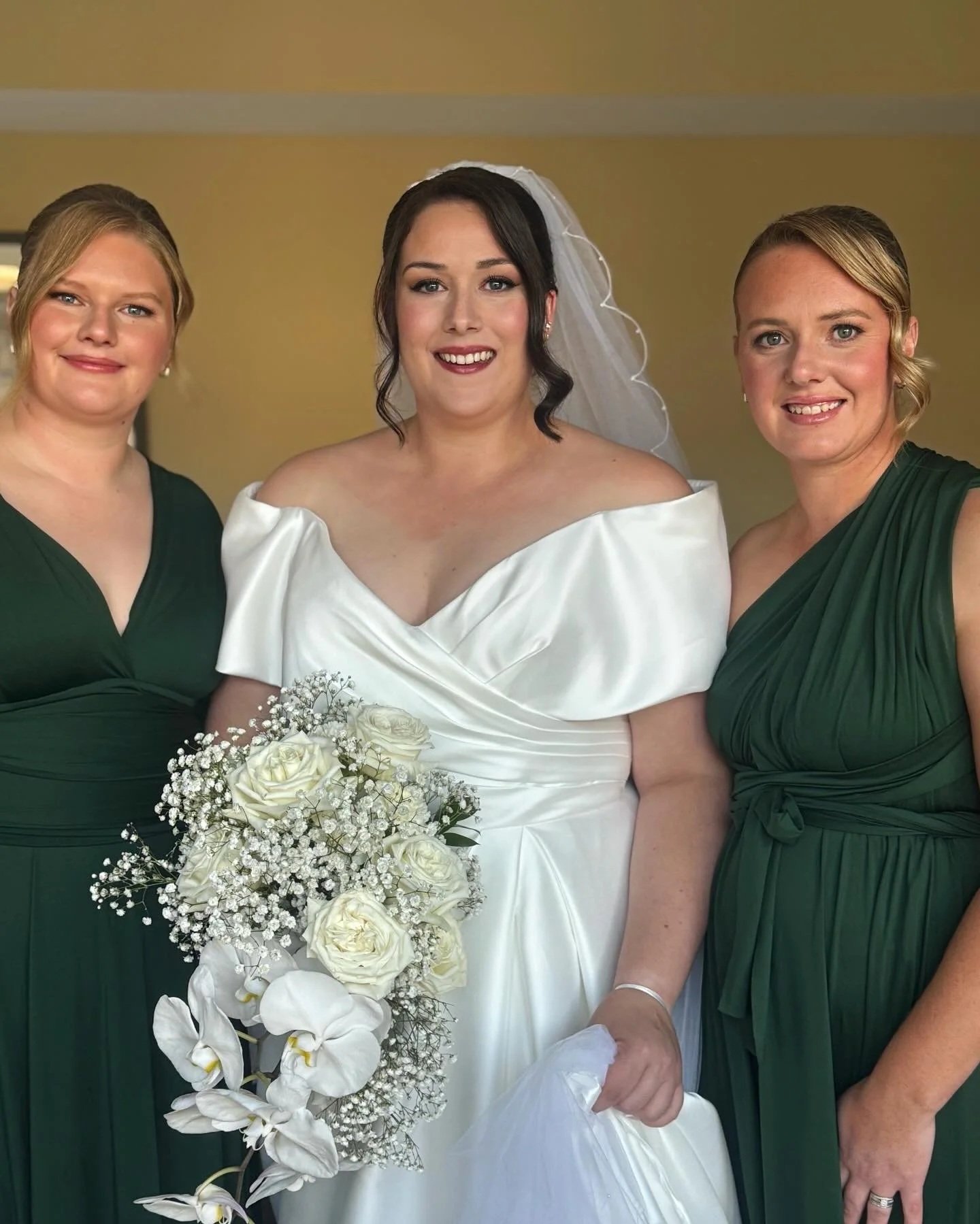 Bride in white wedding dress holding a bouquet of white roses and orchids, flanked by two bridesmaids in green dresses.