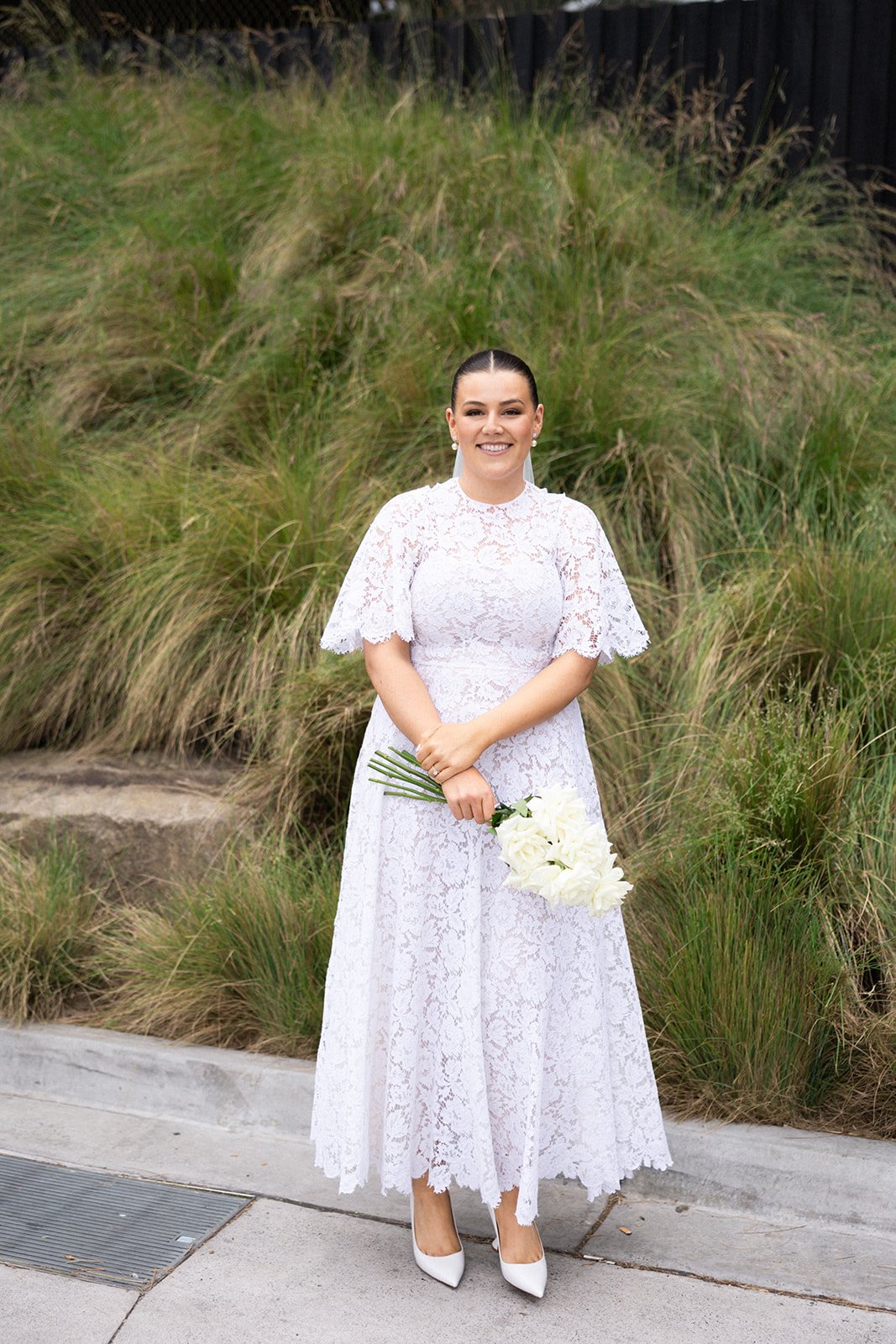 A bride in a white lace dress holding a bouquet of white flowers, standing outdoors in front of green grass and plants, smiling at the camera.