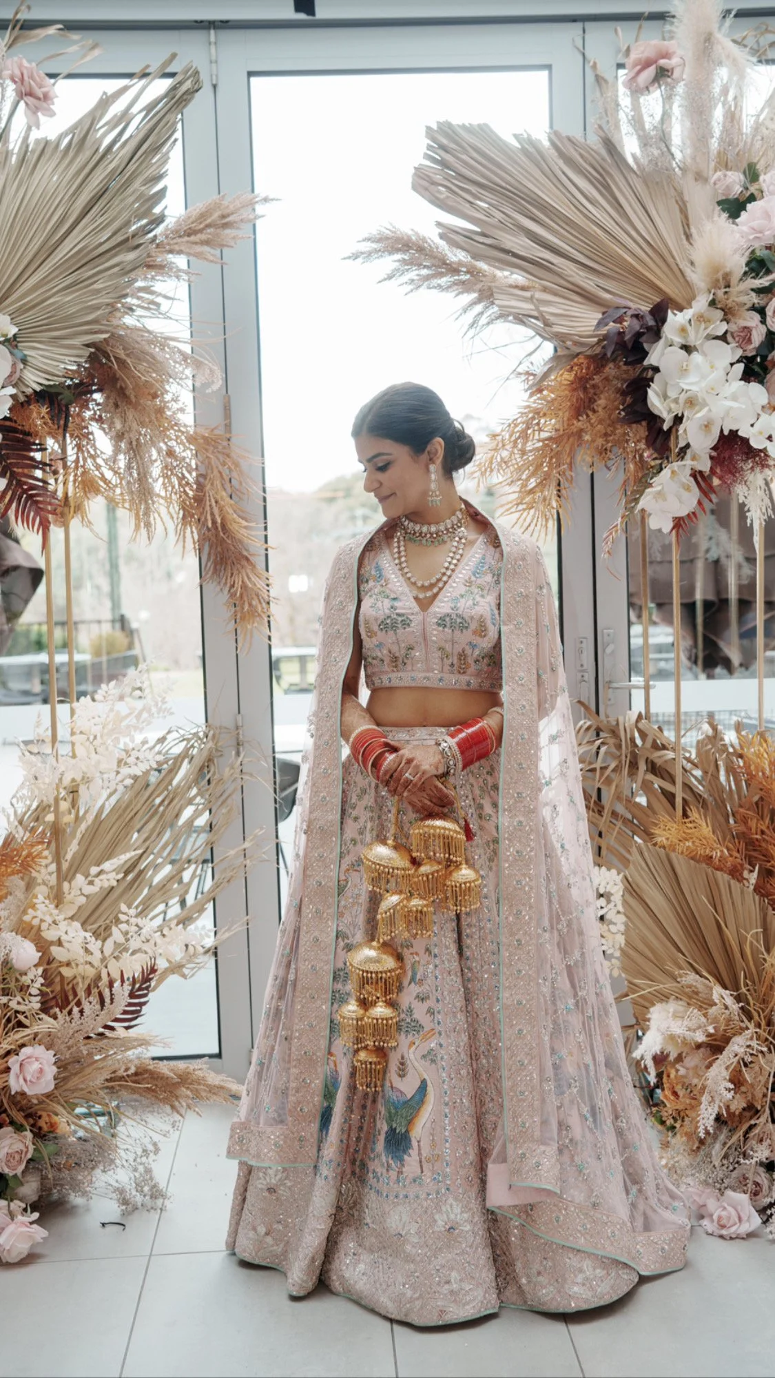 A woman dressed in traditional Indian wedding attire, holding golden bridal ornaments, standing in front of a floral and foliage decoration near a glass door.