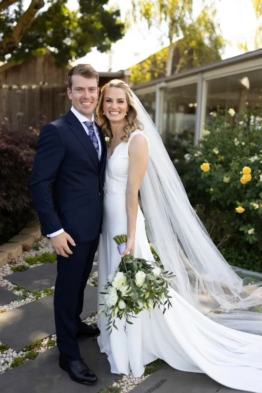 Bride and groom standing outdoors on a wedding day, smiling, with the bride holding a bouquet of white flowers and wearing a white wedding dress with a long veil.