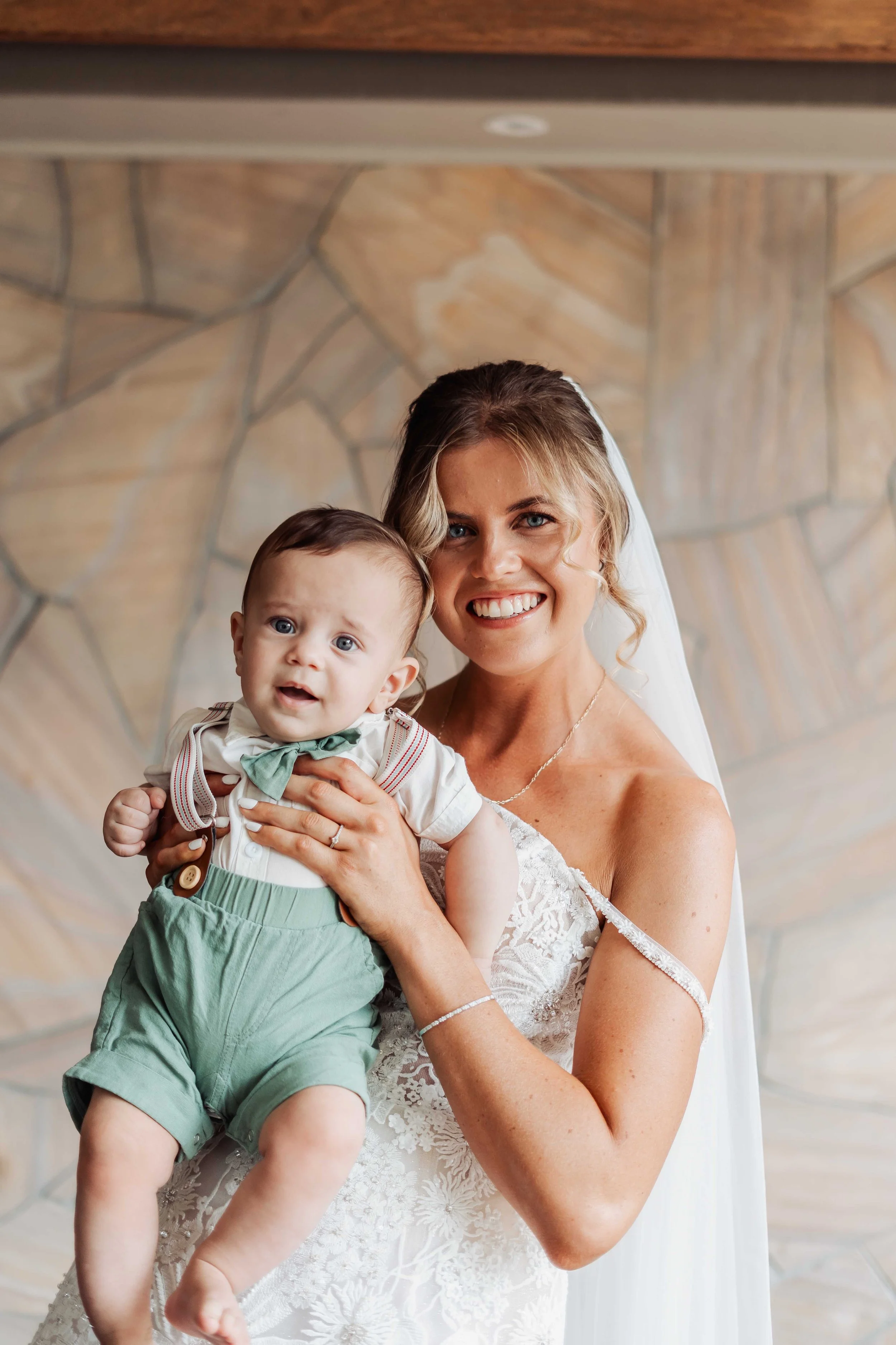 A woman in a wedding dress holding a young boy dressed in green shorts and a cream shirt with a green bow tie.
