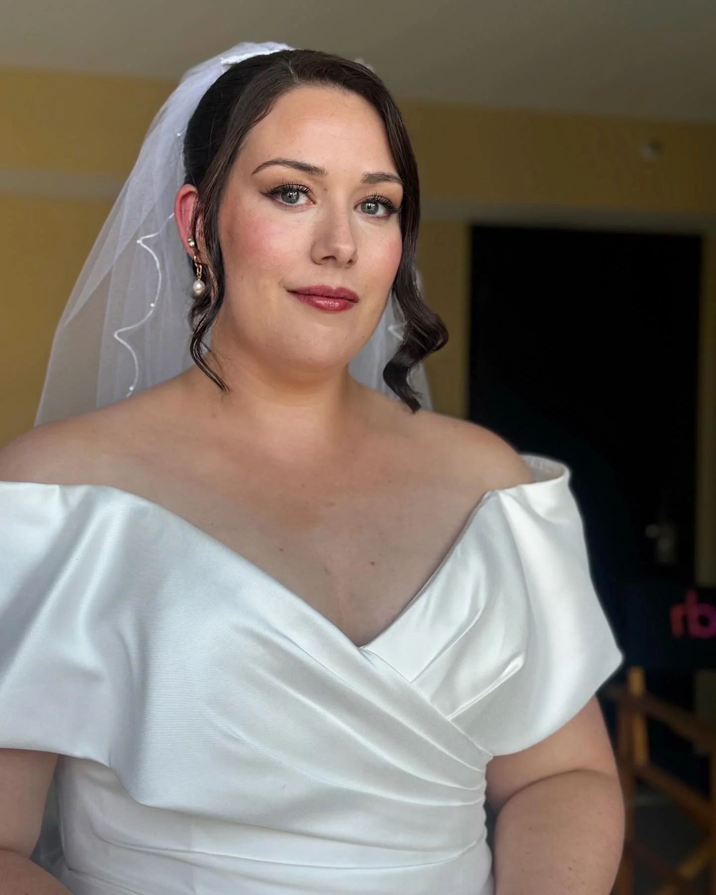 A woman dressed in a wedding gown with a white veil and pearl earrings, standing inside a room with yellow walls.