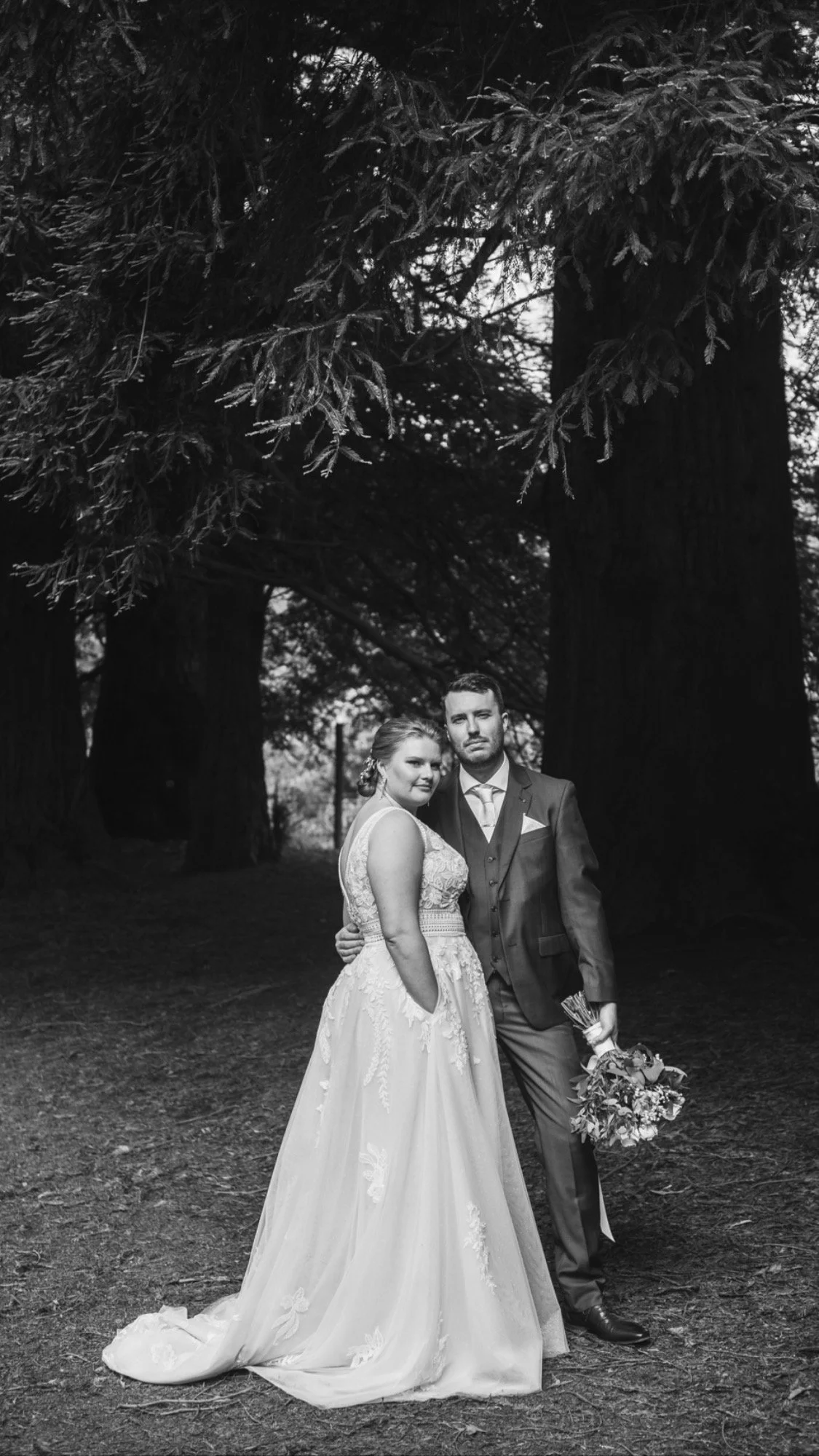 A black and white photo of a bride and groom standing outdoors among large trees, with the bride in a wedding gown and the groom in a suit holding a bouquet.