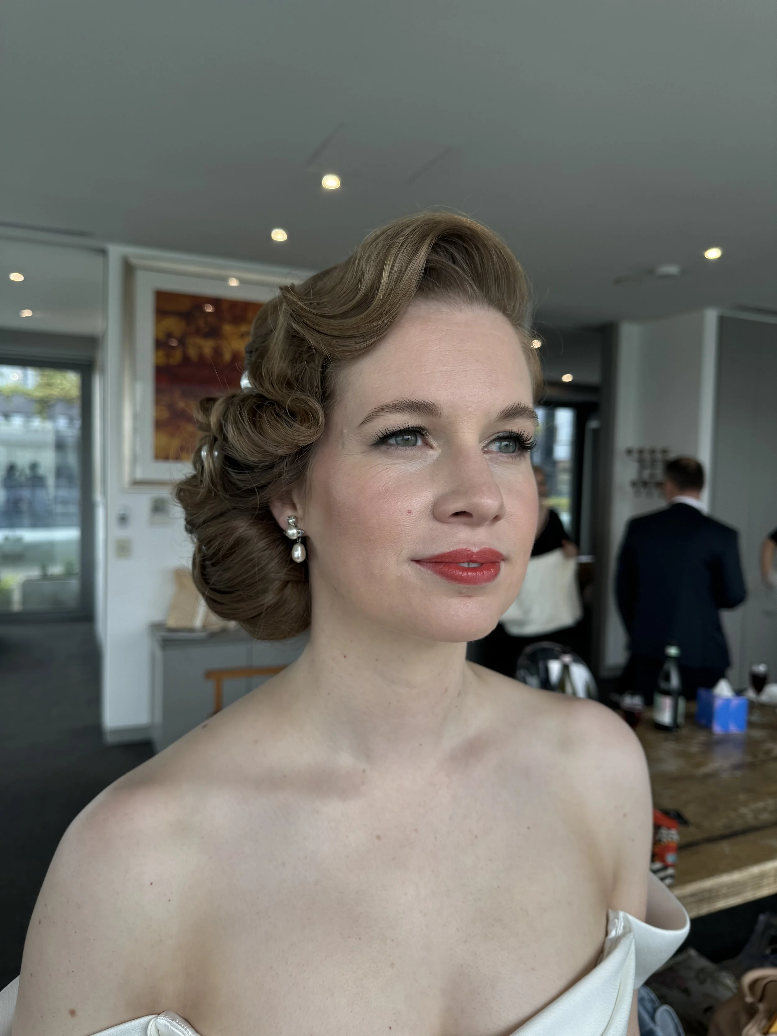 A woman with vintage-style curled hair and red lipstick, wearing pearl earrings, is posing indoors during a formal event.