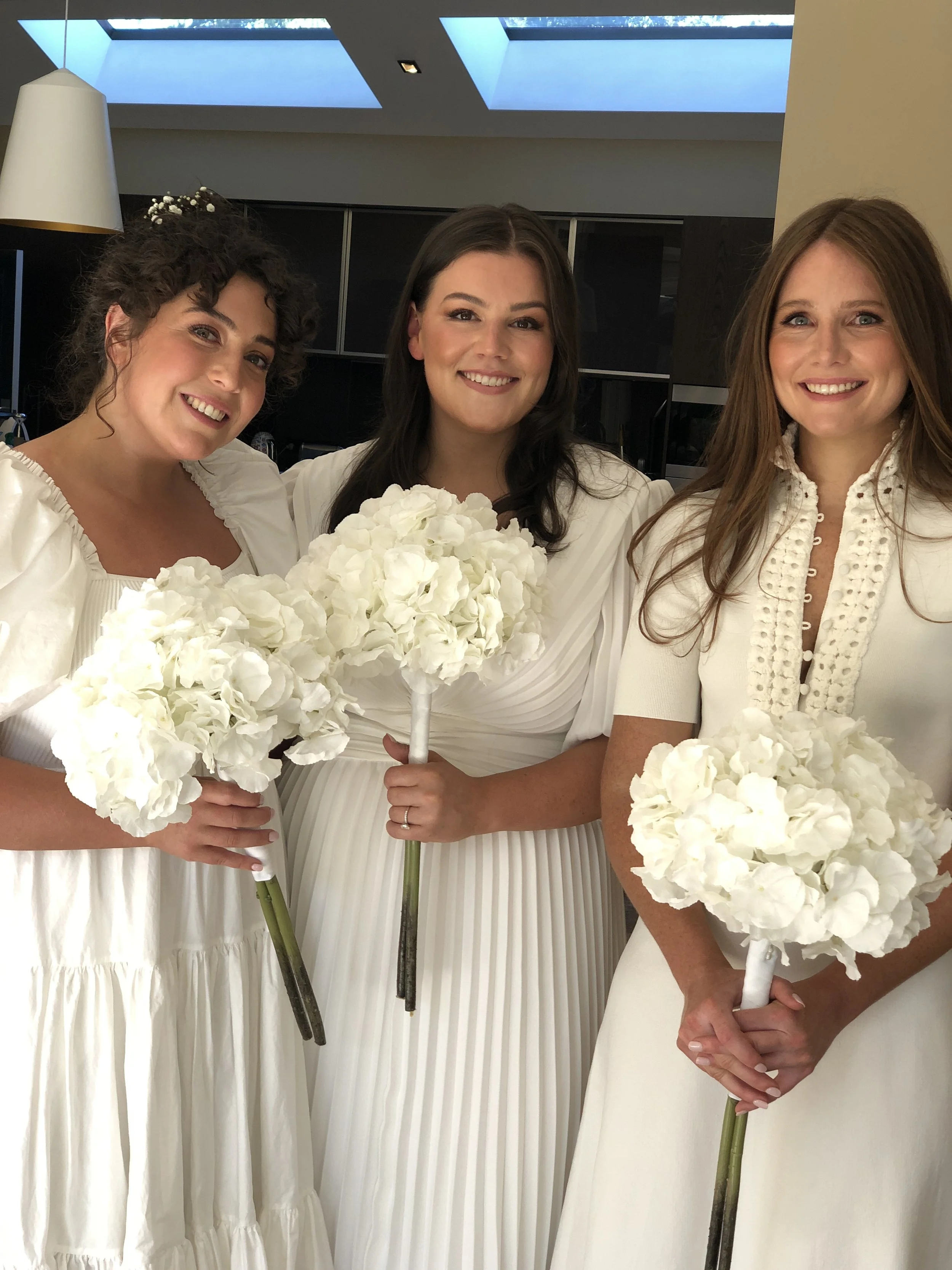 Three women in white dresses holding white flower bouquets, smiling indoors with a modern ceiling and skylights.