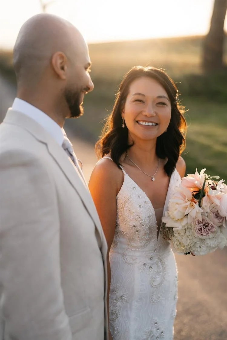 A smiling bride holding a bouquet of flowers on her wedding day, standing outdoors with her groom during sunset.