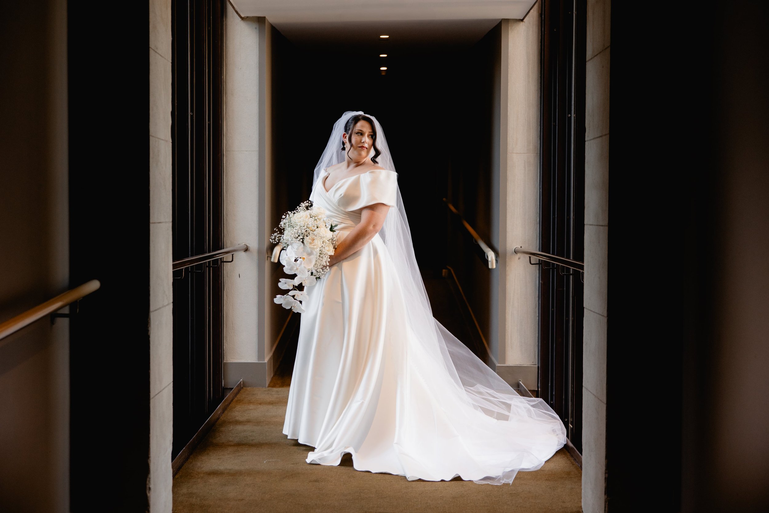 A bride in a white wedding gown with a veil holding a bouquet of white flowers, standing in an elevator at her wedding in Melbourne.
