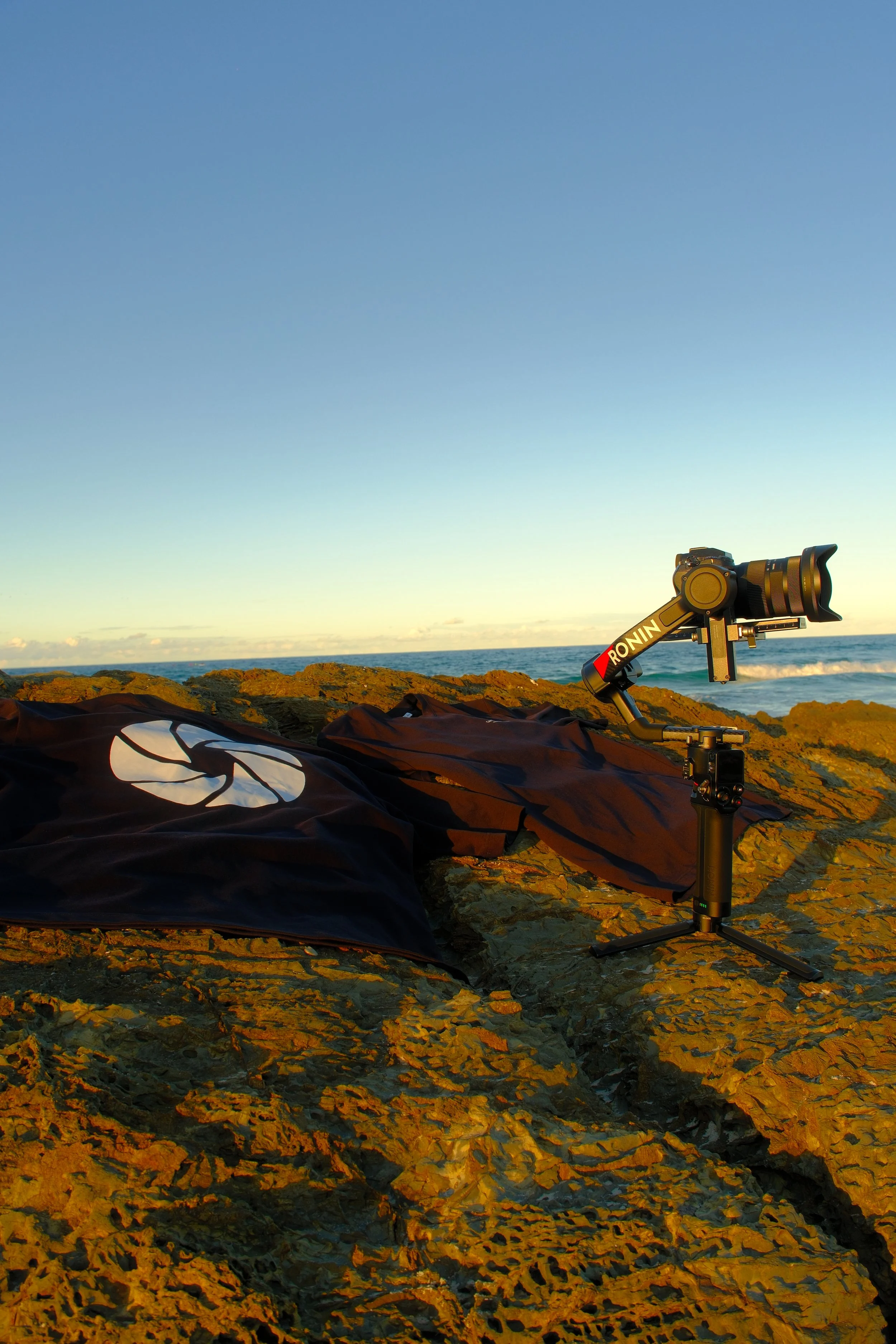 A camera mounted on a tripod facing the ocean, with a drone or kite on rocky terrain during sunset or sunrise.