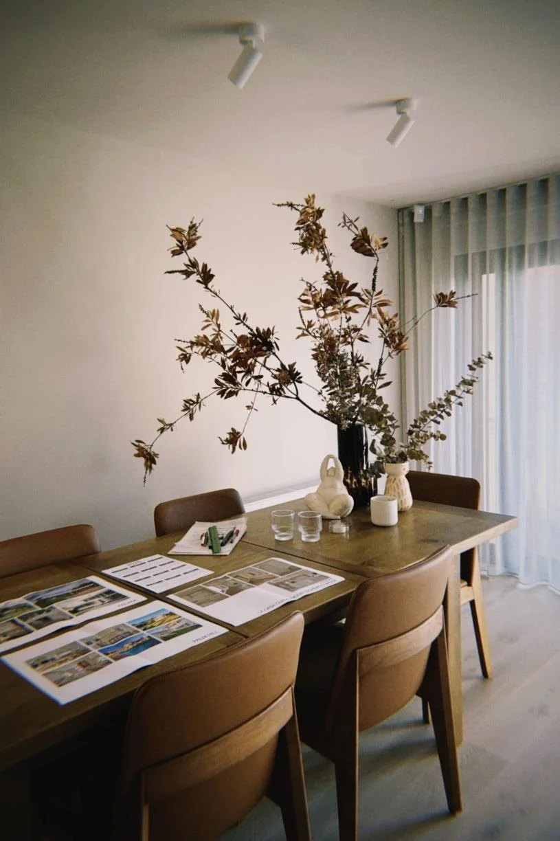 Dining table with chairs, large vase with dried branches, decorative items, glasses, and open magazines or catalogs, next to a window with sheer white curtains.