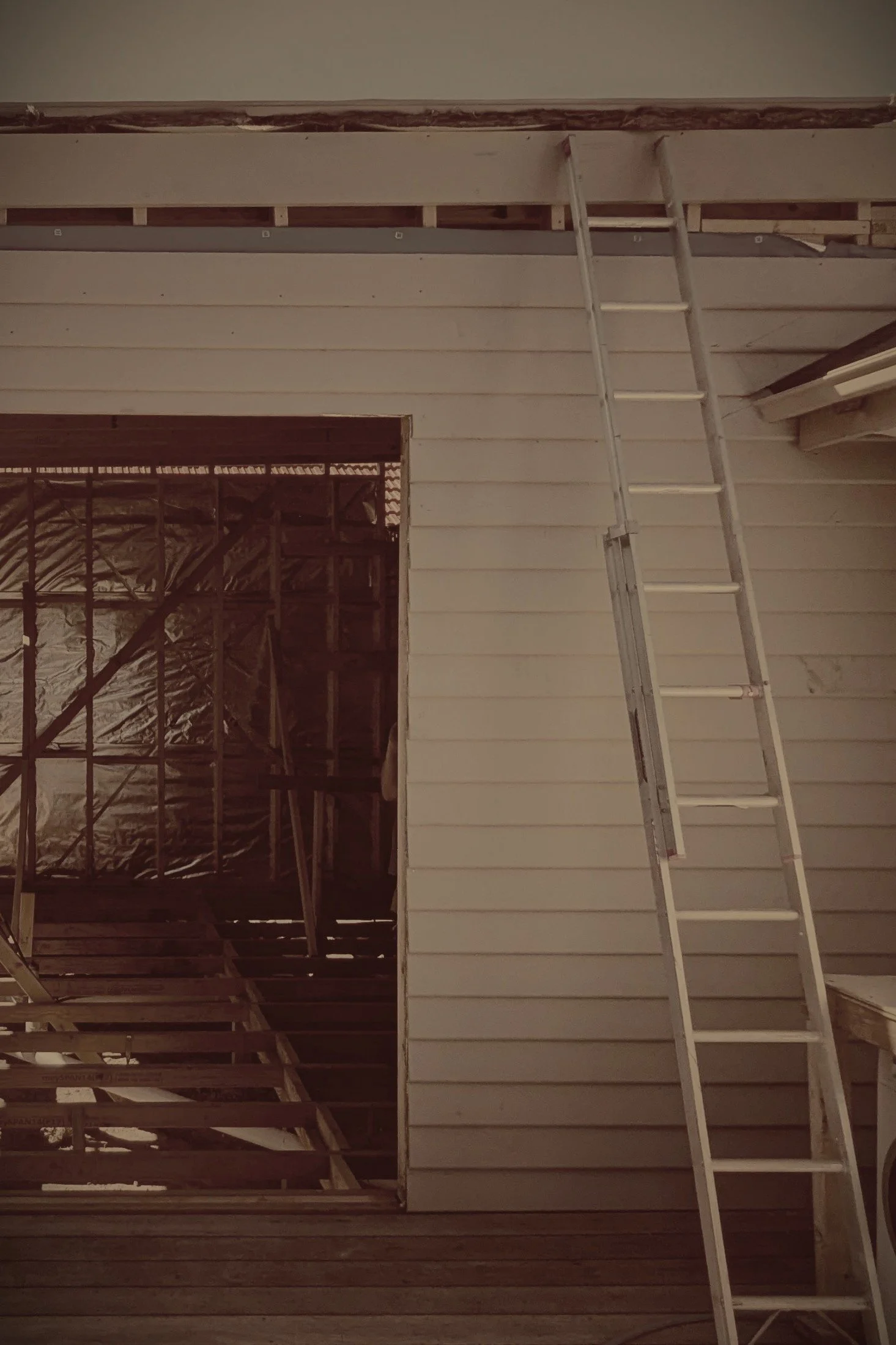 Interior of a house under renovation with a partially completed second story, a ladder leaning against the wall, and visible wooden framing and insulation.