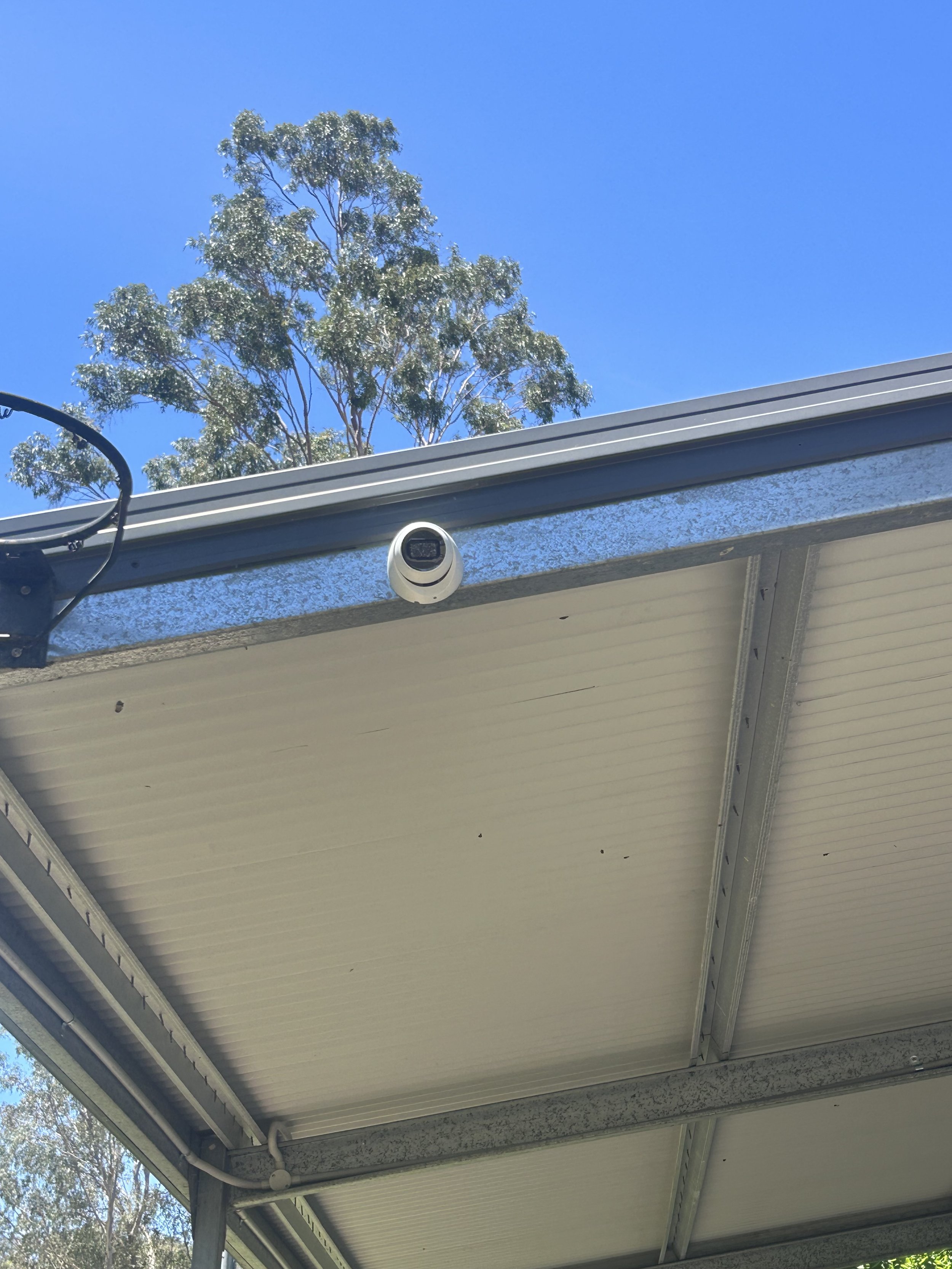 View of the underside of a house roof with a security camera attached, some trees in the background, and a clear blue sky.