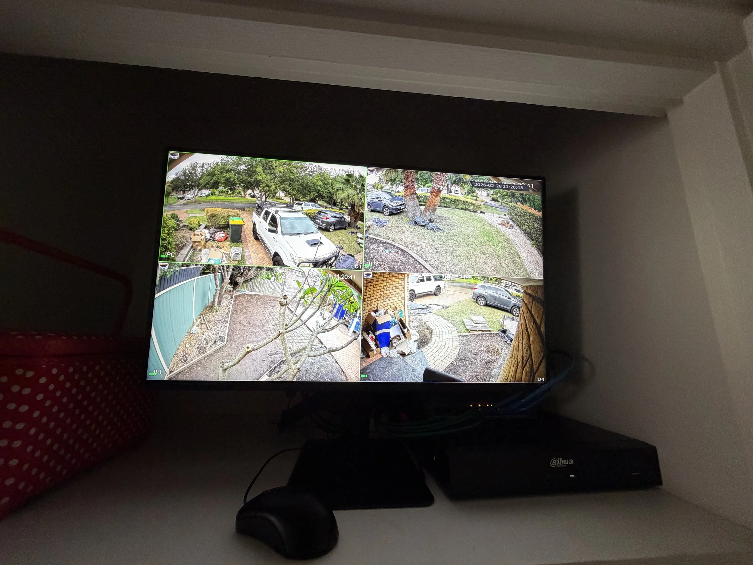 Security camera monitor displaying four surveillance camera views of a residential area with cars, trees, and yard.