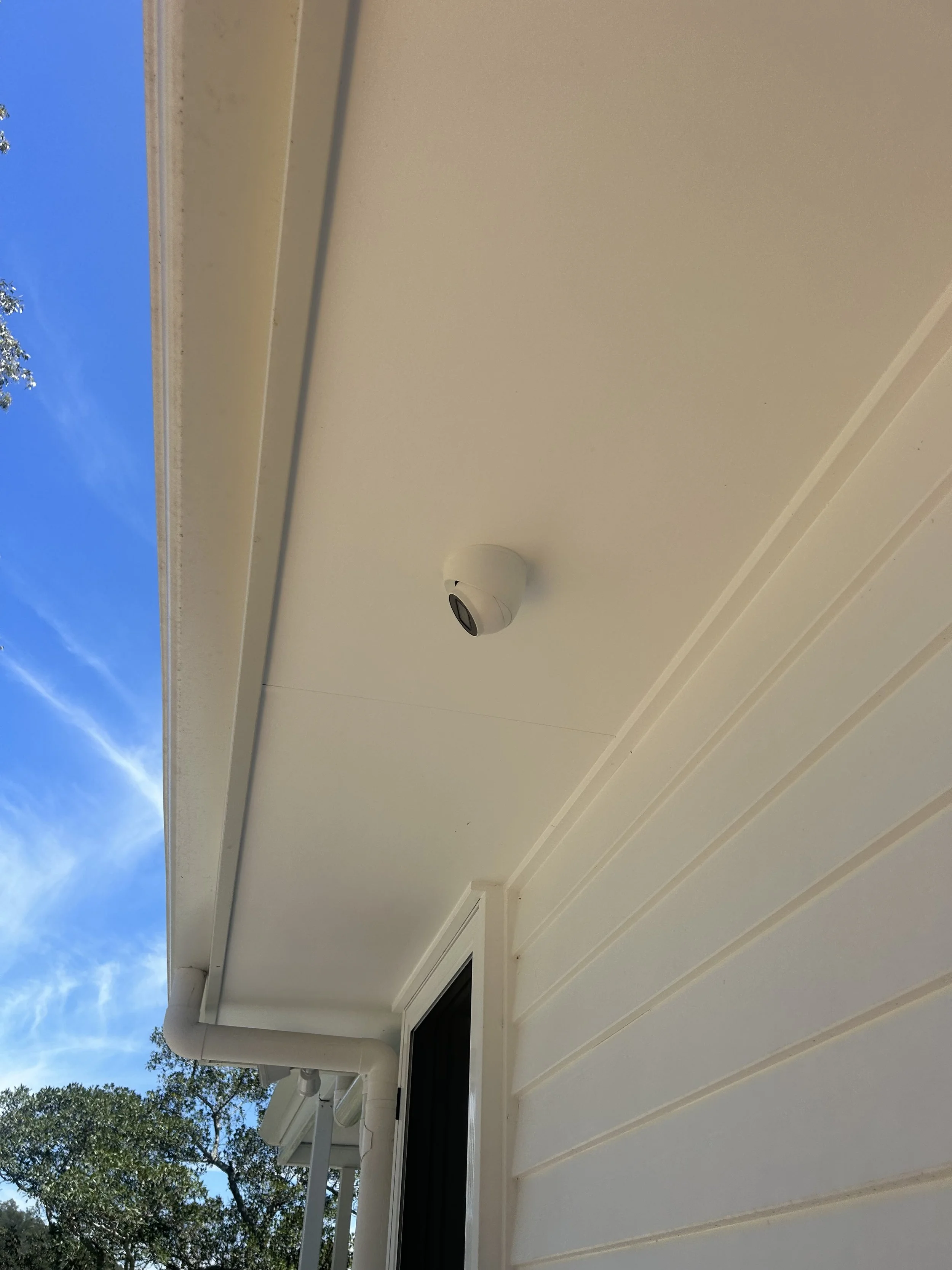 View of a beige house exterior with a small window, white gutter, downspout, and a round outdoor light fixture. Part of a leafy tree is visible in the background against a blue sky.