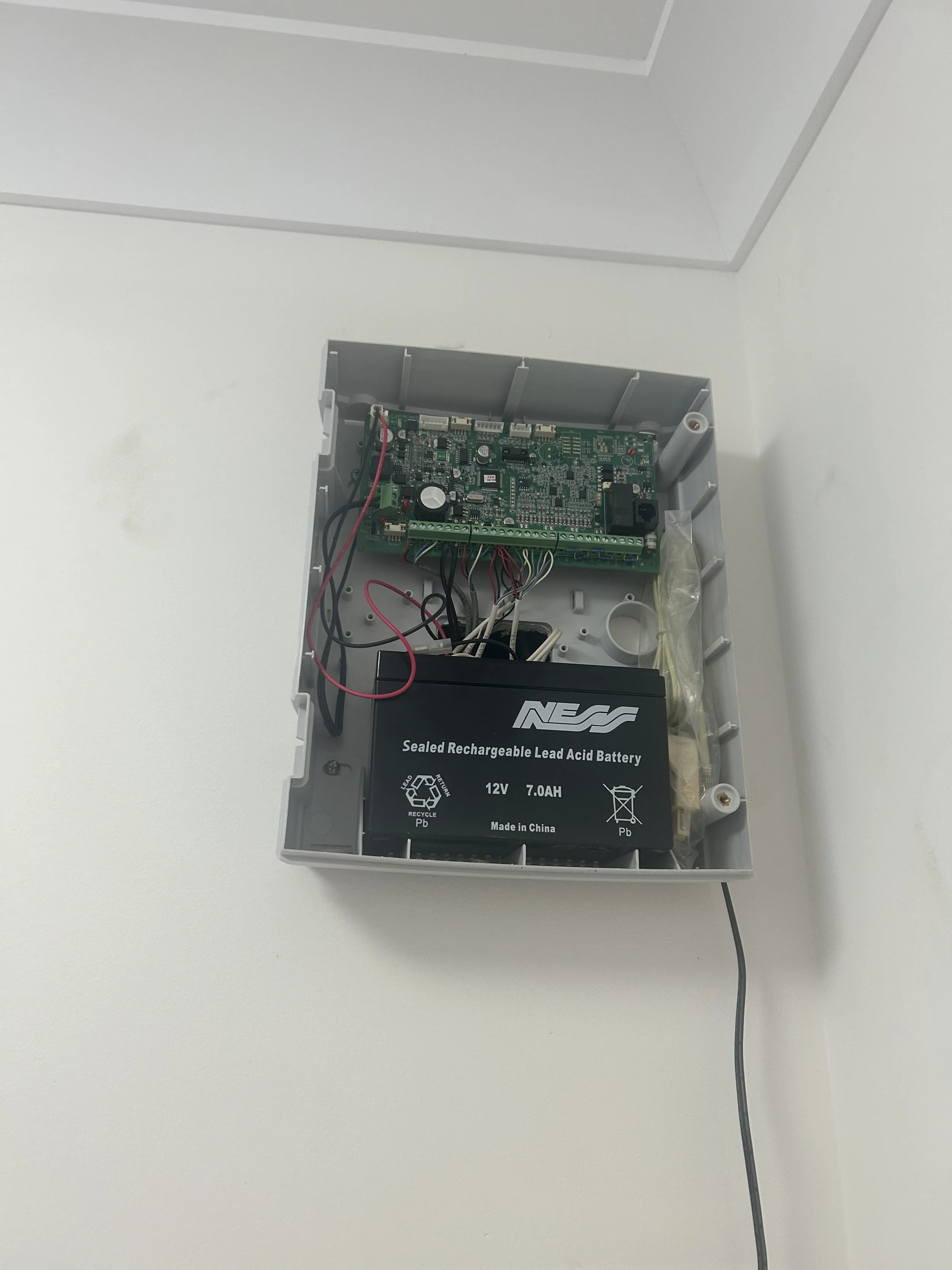 Open electrical box with circuit board, wires, and a battery inside a white wall-mounted enclosure.