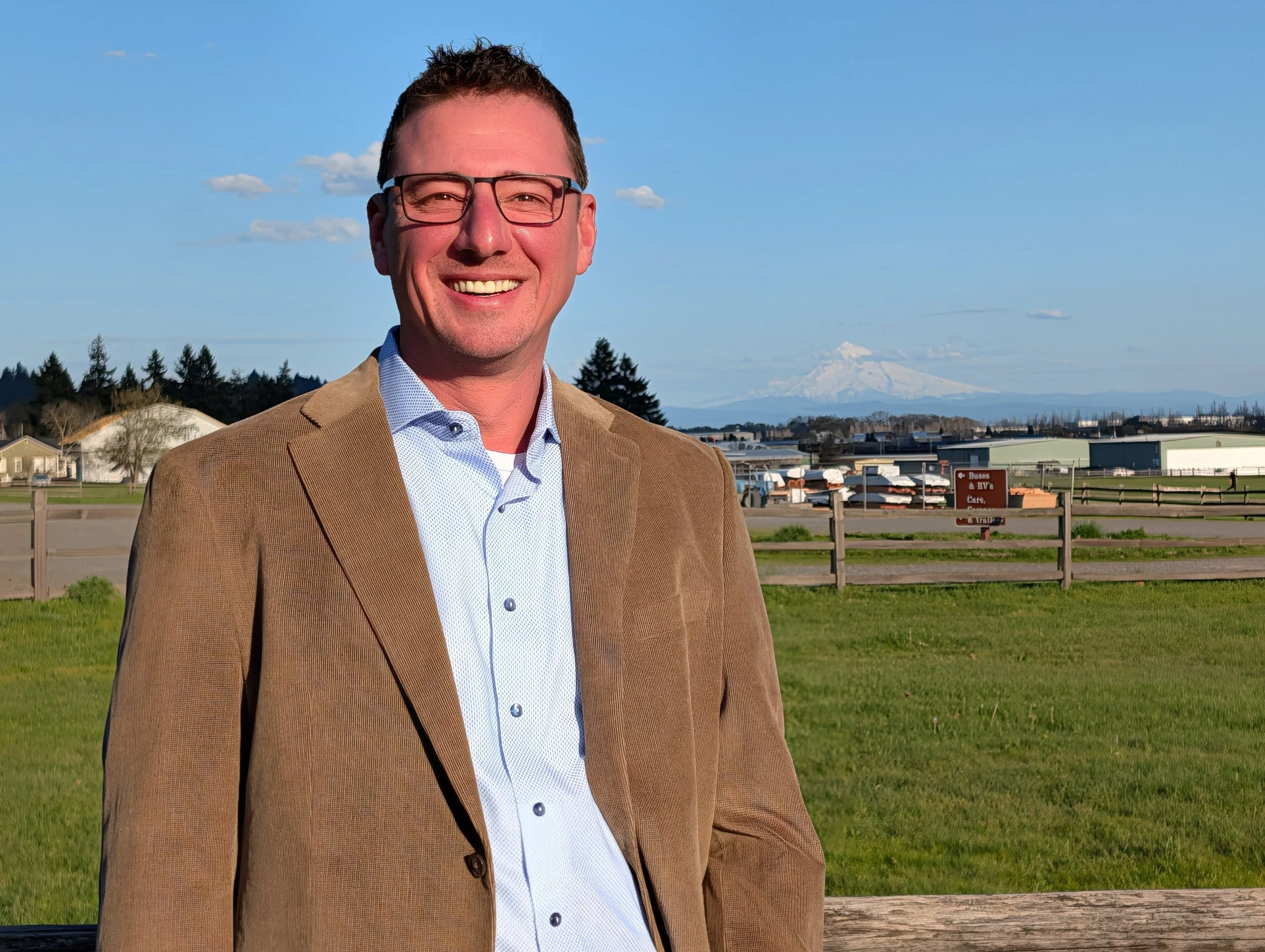 A smiling Lukas Bardue, candidate for Clark County Council District 1 in glasses and a tan blazer standing outdoors at Fort Vancouver with green grass, a wooden fence, Pearson Air Museum, and snow-capped Mt Hood in the background.