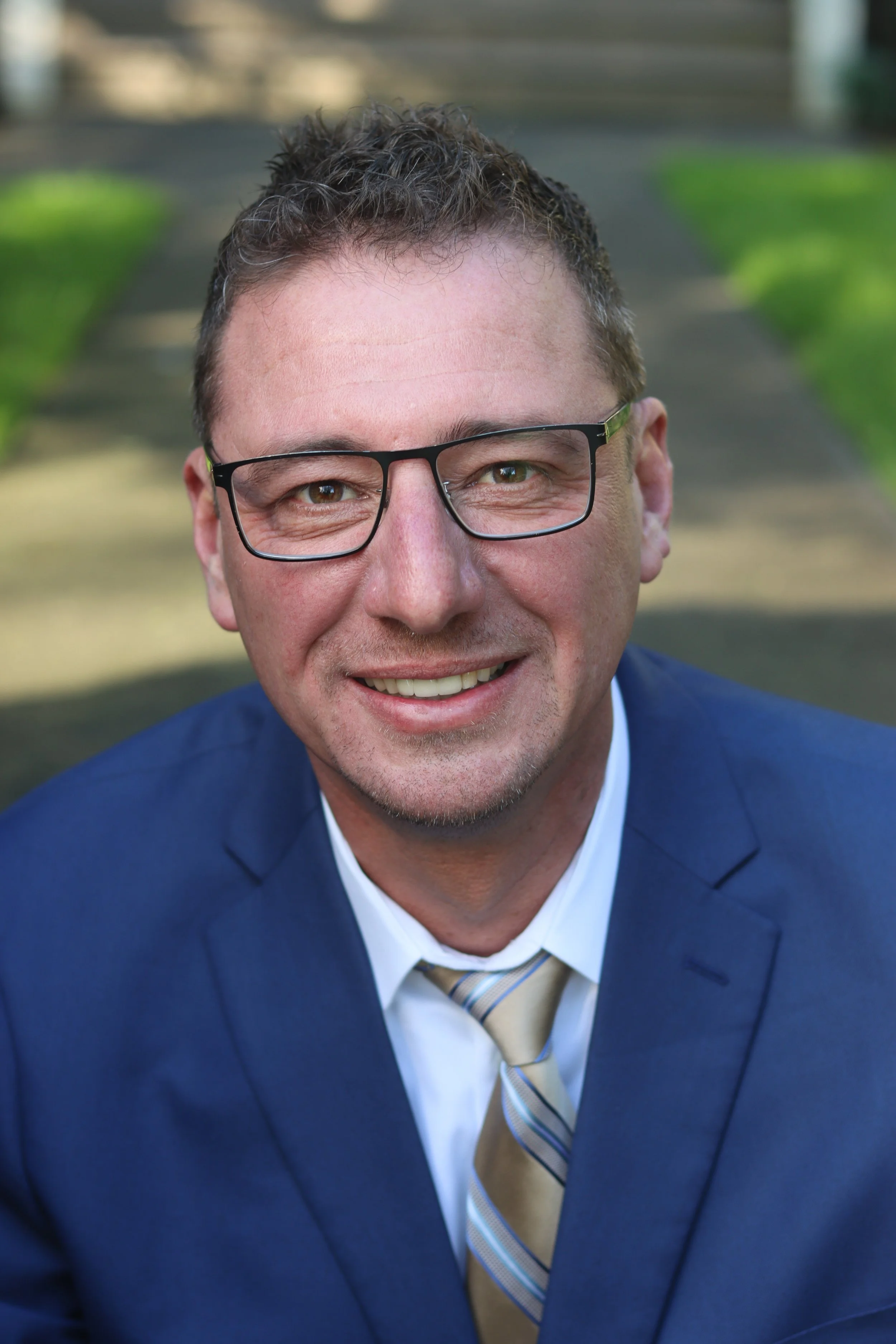A smiling Lukas Bardue, candidate for Clark for Clark County Council District 1,  in a navy blue suit, white shirt, and striped tie outdoors with a walkway and grass in the background.