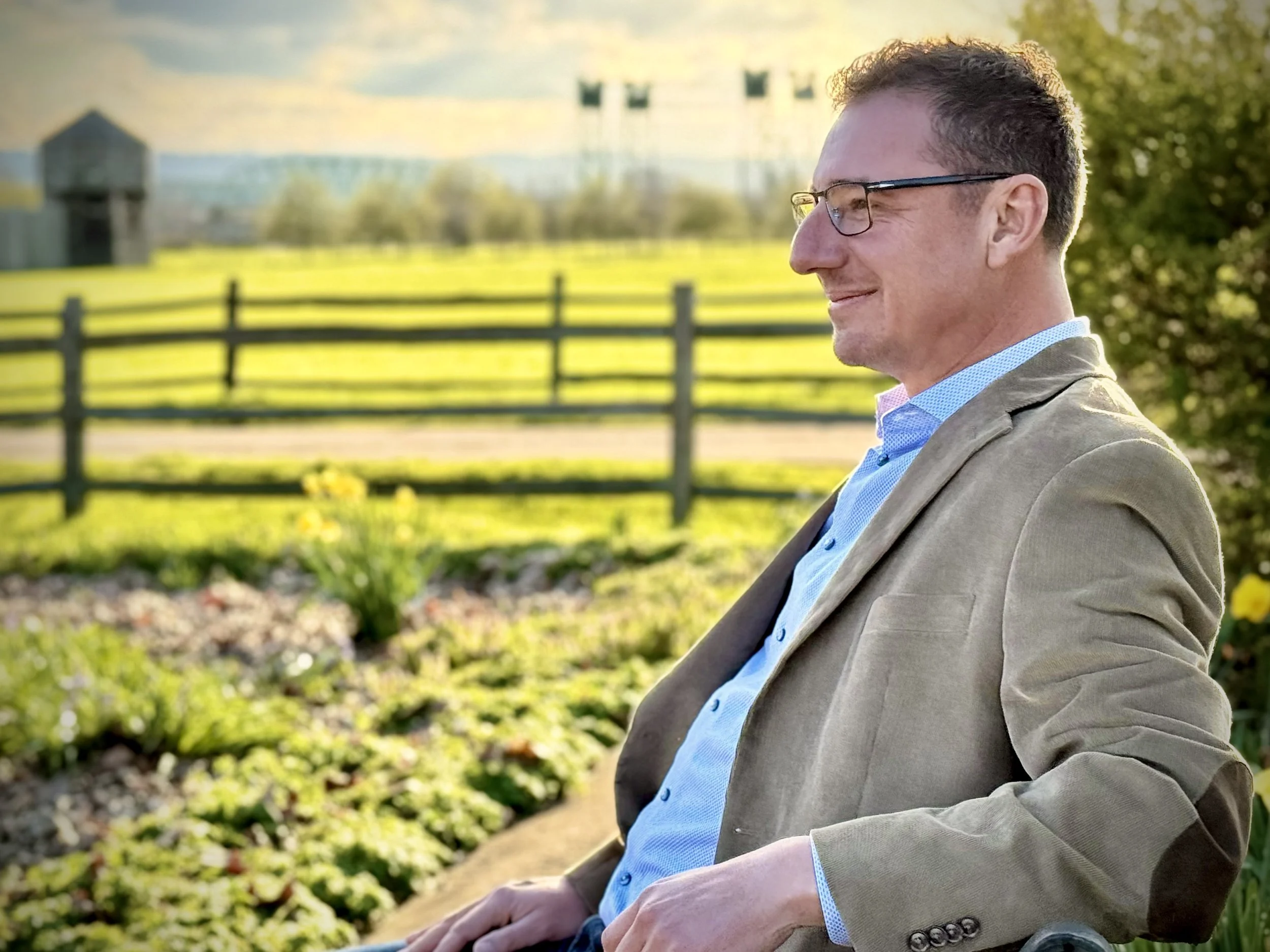 Lukas Bardue, candidate for Clark for Clark County Council District 1, sitting outdoors in Fort Vancouver garden during daytime, wearing a beige blazer and a blue shirt, with a wooden fence, bright yellow flowers, and I-5 bridge in the background.