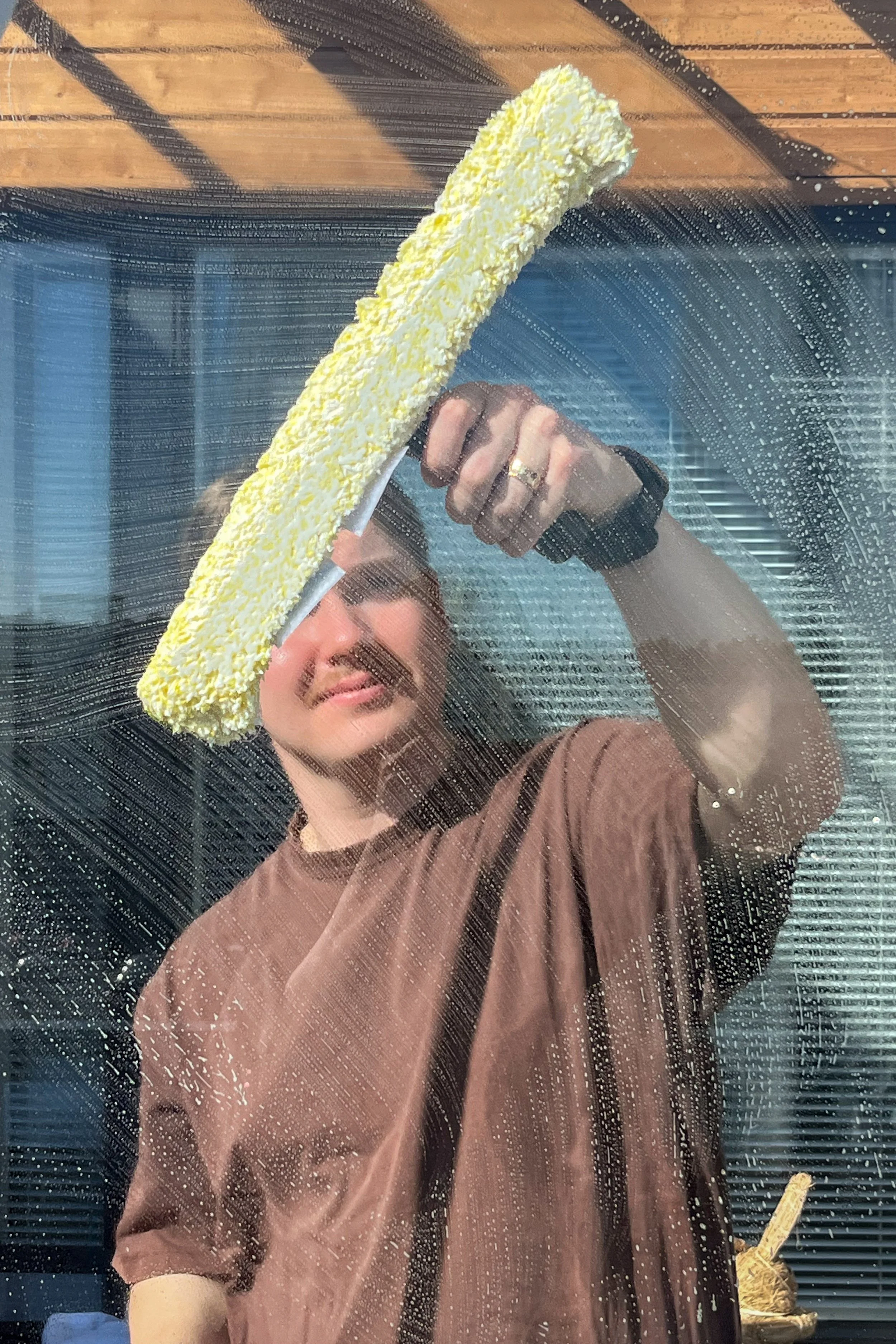 Person cleaning a glass window with a yellow sponge, visible streaks of soap and cleaning spray.