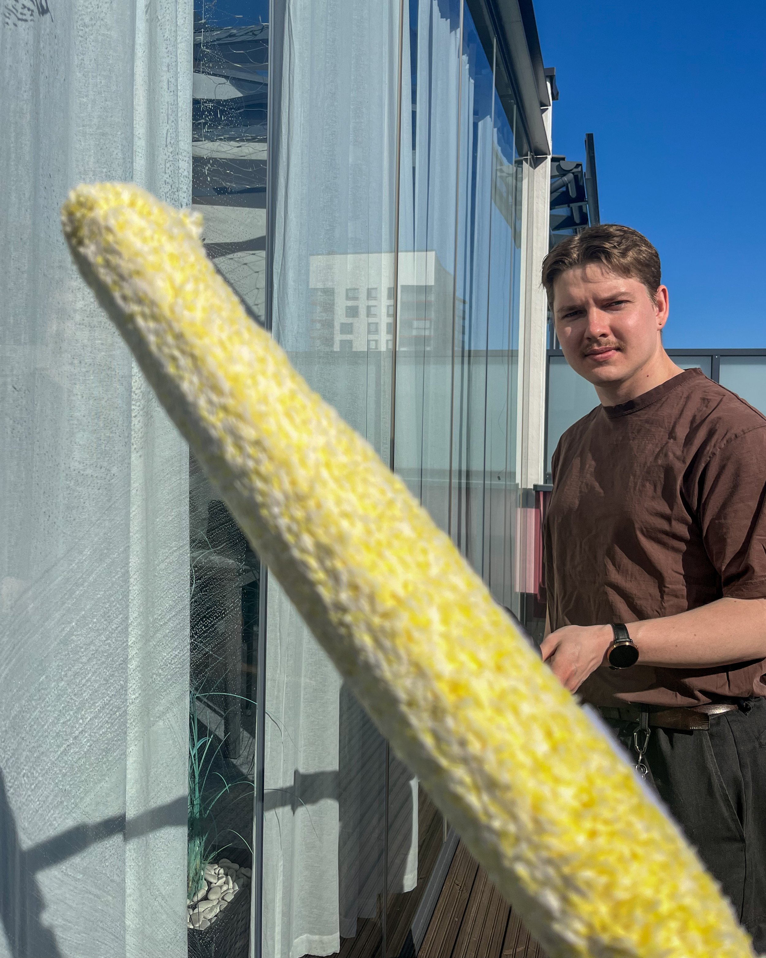 A young man with brown hair and a mustache stands outside on a balcony, wearing a brown shirt and black watch, looking at the camera. In the foreground, a yellow and white object obstructs part of the view.
