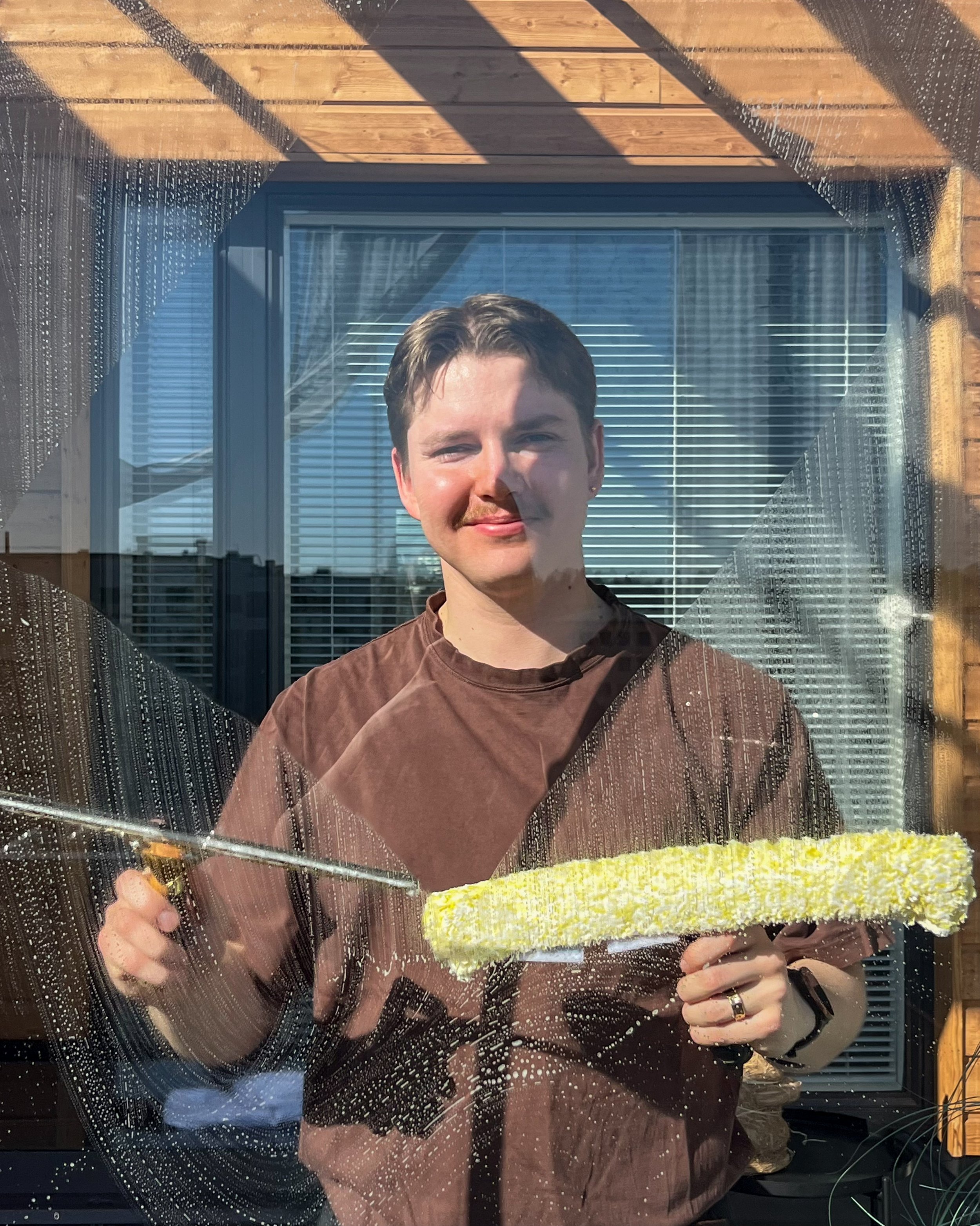 A young man cleaning a glass window with a yellow sponge, sunlight shining on his face, a wooden frame around the window, and blinds visible through the glass.