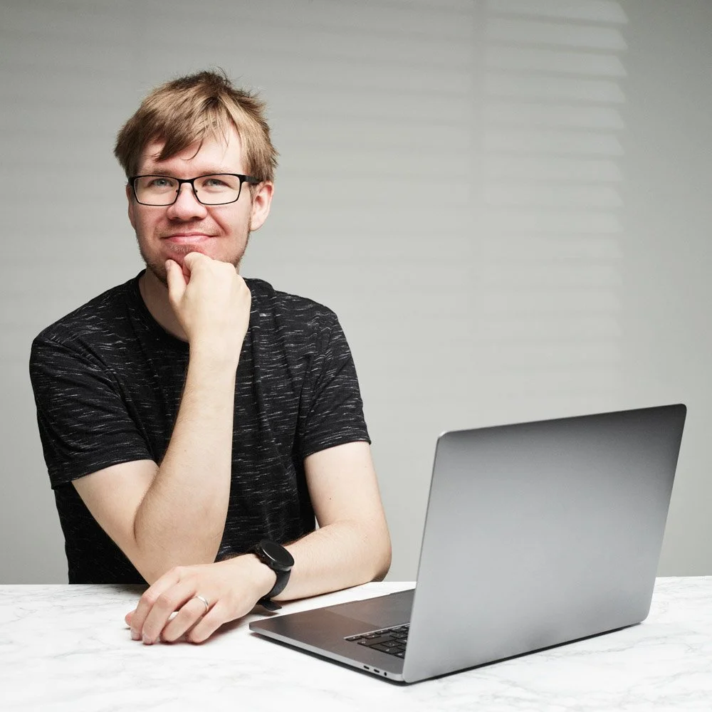 A young man with glasses, light brown hair, and a black t-shirt sitting at a white table with a silver laptop, resting his chin on his left hand and smiling.