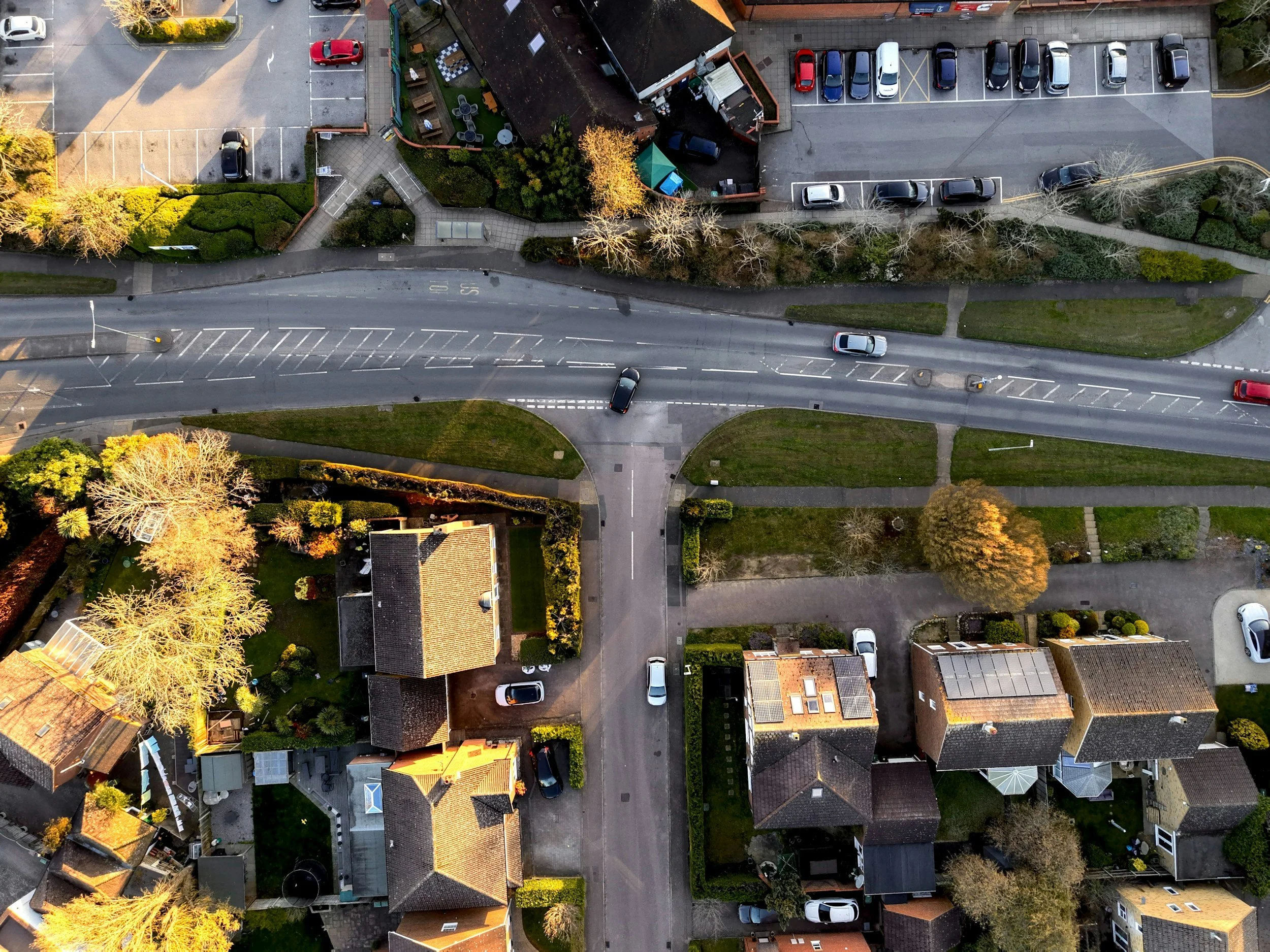 An aerial view of a residential neighborhood shows houses, trees, streets, parked and moving cars, and a parking lot. The houses have yards with grass and bushes, with some having solar panels on roofs. The streets are lined with trees and sidewalks.
